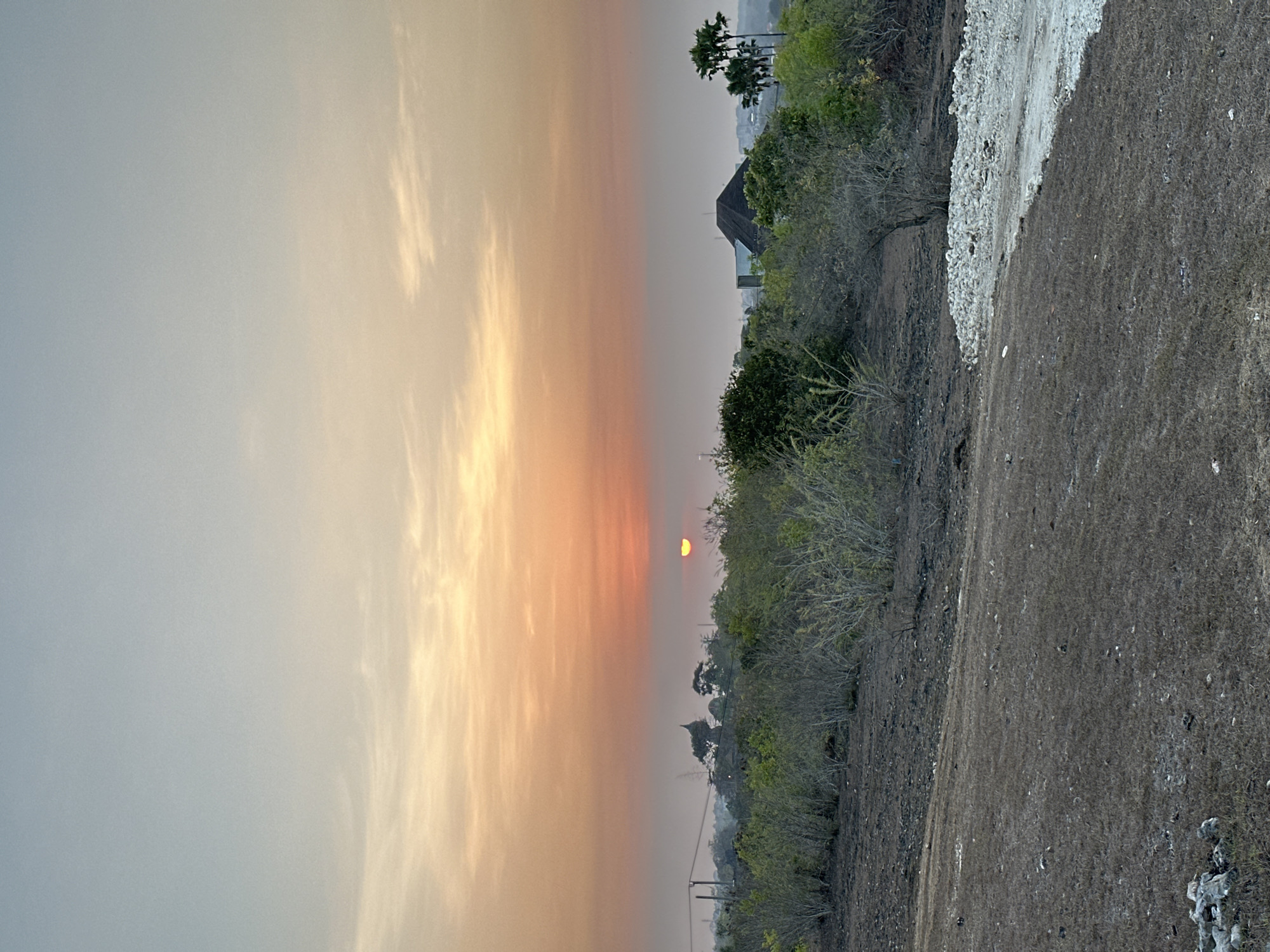A sunset is captured over a semi-arid landscape in Kuta Selatan, Indonesia. The sky transitions from a soft blue at the top to a gradient of pale orange and deep orange hues near the horizon. A sliver of the sun is visible as a bright orange disk, partially obscured by the horizon line. In the middle ground, a line of dense vegetation, primarily consisting of light green bushes and trees, stretches across the frame. Beyond the foliage, structures are faintly visible, including a building with a dark, pitched roof and what appears to be a domed structure topped with a finial. Several palm trees stand to the right of the frame, silhouetted against the sky. The foreground is a textured expanse of dry, dark brown earth interspersed with sparse, dry grass and scattered small stones. A lighter, gravelly path or pile of stones is present in the lower right quadrant of the image. There are no people or animals visible in the scene.