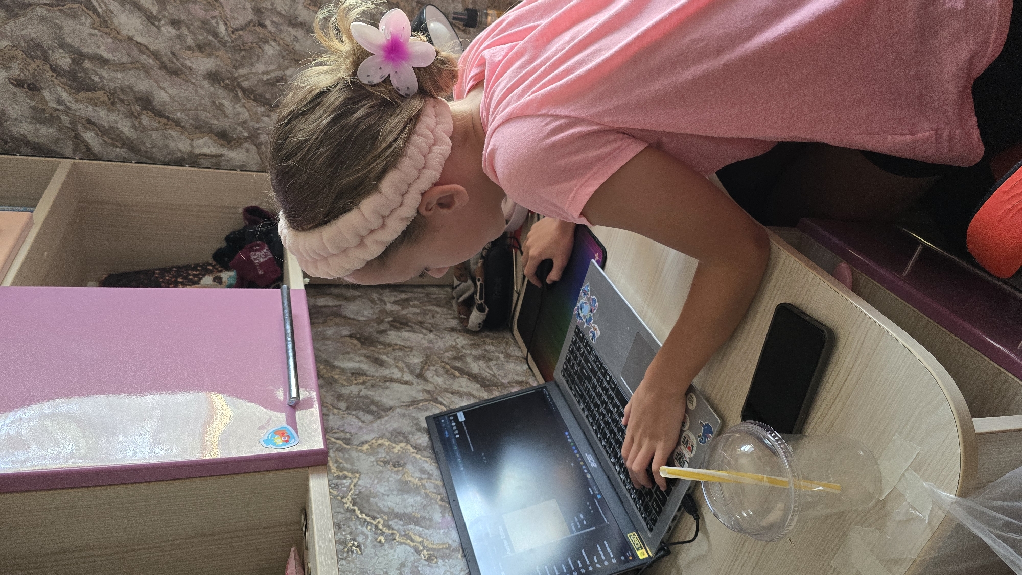 A young girl with blonde hair tied up with a pink headband and a pink flower accessory, wearing a pink t-shirt, is sitting at a desk with a laptop. She is looking at the laptop screen and her right hand is on the mouse. A clear plastic cup with a yellow straw is on the desk next to the laptop, along with a black smartphone. The desk is made of light-colored wood with a marble-patterned wallpaper behind it. There is a pink cabinet above the desk and shelves to the right. The setting appears to be an indoor room, possibly a bedroom or study area in Novohuivynske, Ukraine.