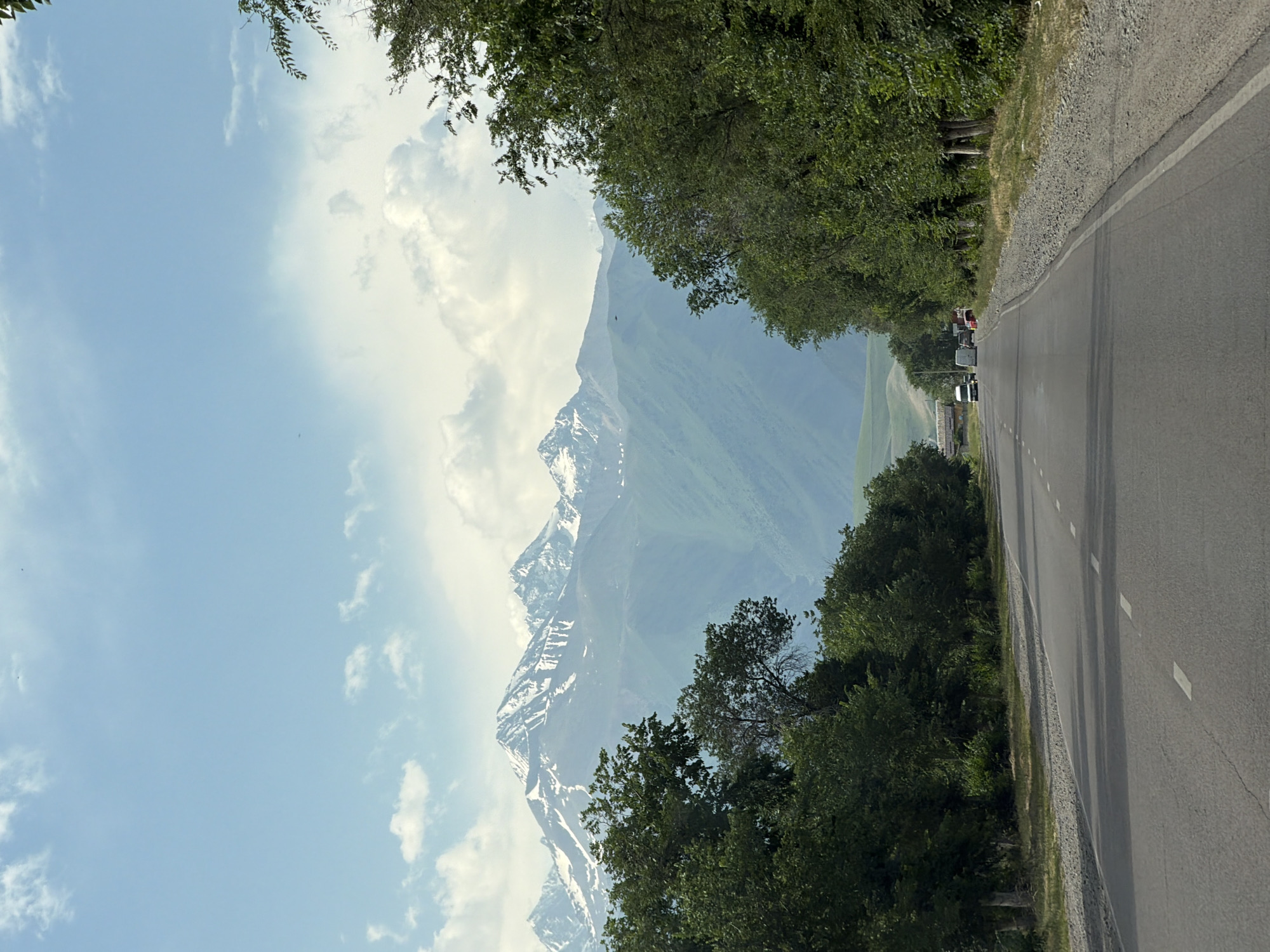 The image captures a scenic view of a road leading towards a majestic mountain range. The road, with a white dashed line marking its lanes, is flanked by lush green trees on both sides. In the distance, snow-capped mountains rise against a backdrop of a bright blue sky with scattered white clouds. Several vehicles, including vans and what appears to be a truck, are visible on the road, suggesting it is a route for transportation. The environment appears to be a rural or semi-rural area, possibly a scenic route in a mountainous region.