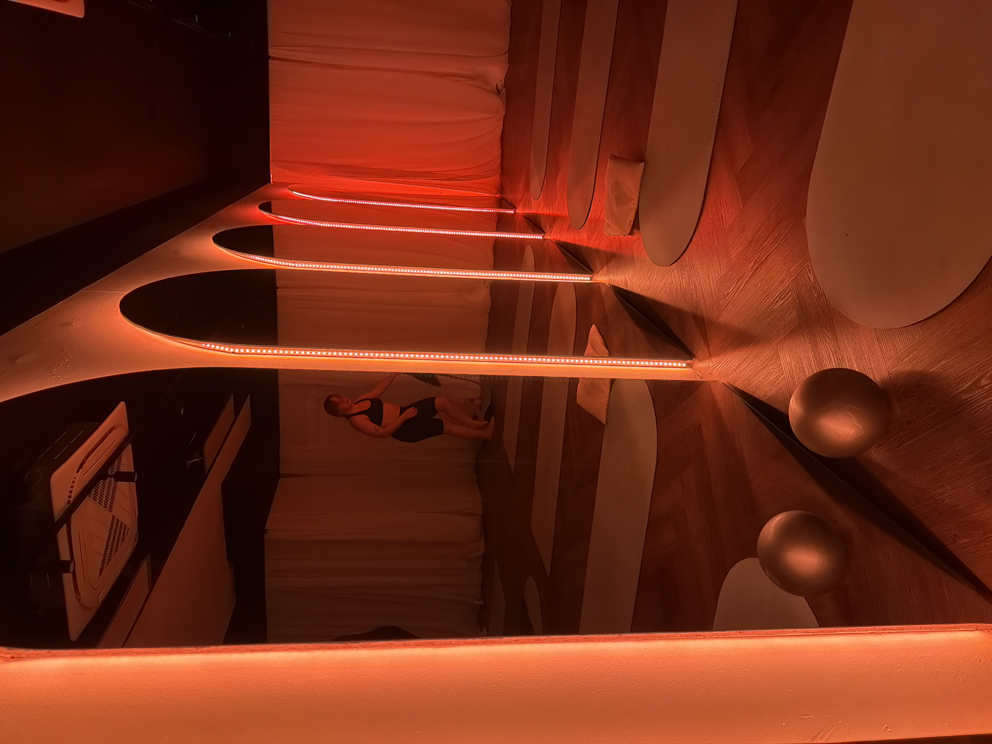 The image depicts a room with a red lighting scheme. The floor is wood with light-colored, oval inlays. Two spherical objects sit on the floor near the viewer's perspective. Overhead, there are several oval mirrors with strip lighting above them. The mirrors reflect a scene with a woman lying on a bed, as well as a ventilation unit. The reflections create a repeating pattern. The location is likely a hotel room, potentially in Moscow, Russia, given the lack of identifying markers and the general design aesthetic.