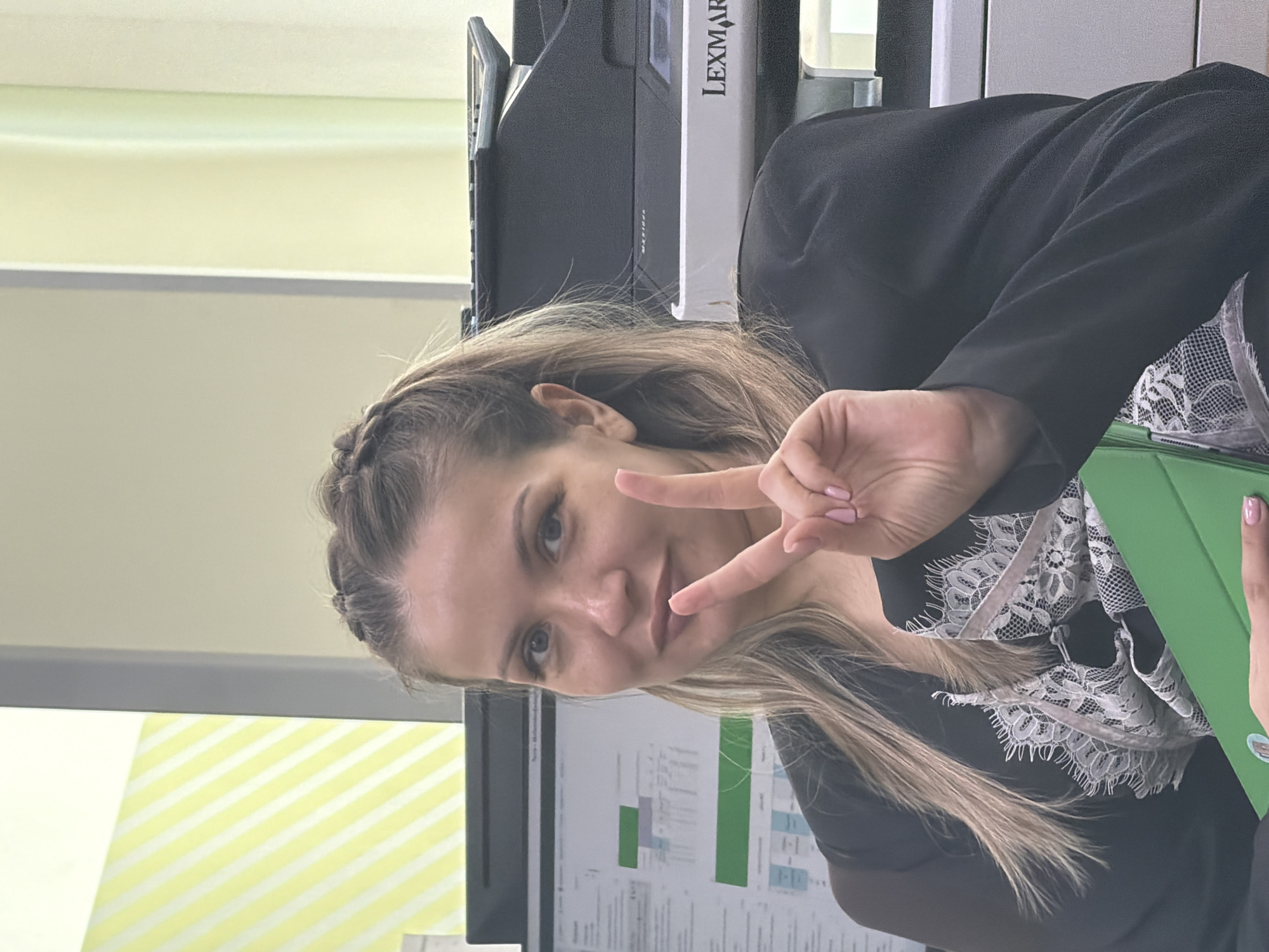 The image shows a woman in a black blazer and a lace-trimmed top, positioned indoors, likely in an office setting. She is making a "peace" sign with her hand, her blonde hair braided at the top and flowing down. Behind her is a computer screen displaying charts and graphs. A Lexmark printer is visible as well. Based on the presence of the local "green" folder, the location is likely within Krasnoyarsk, Russia.