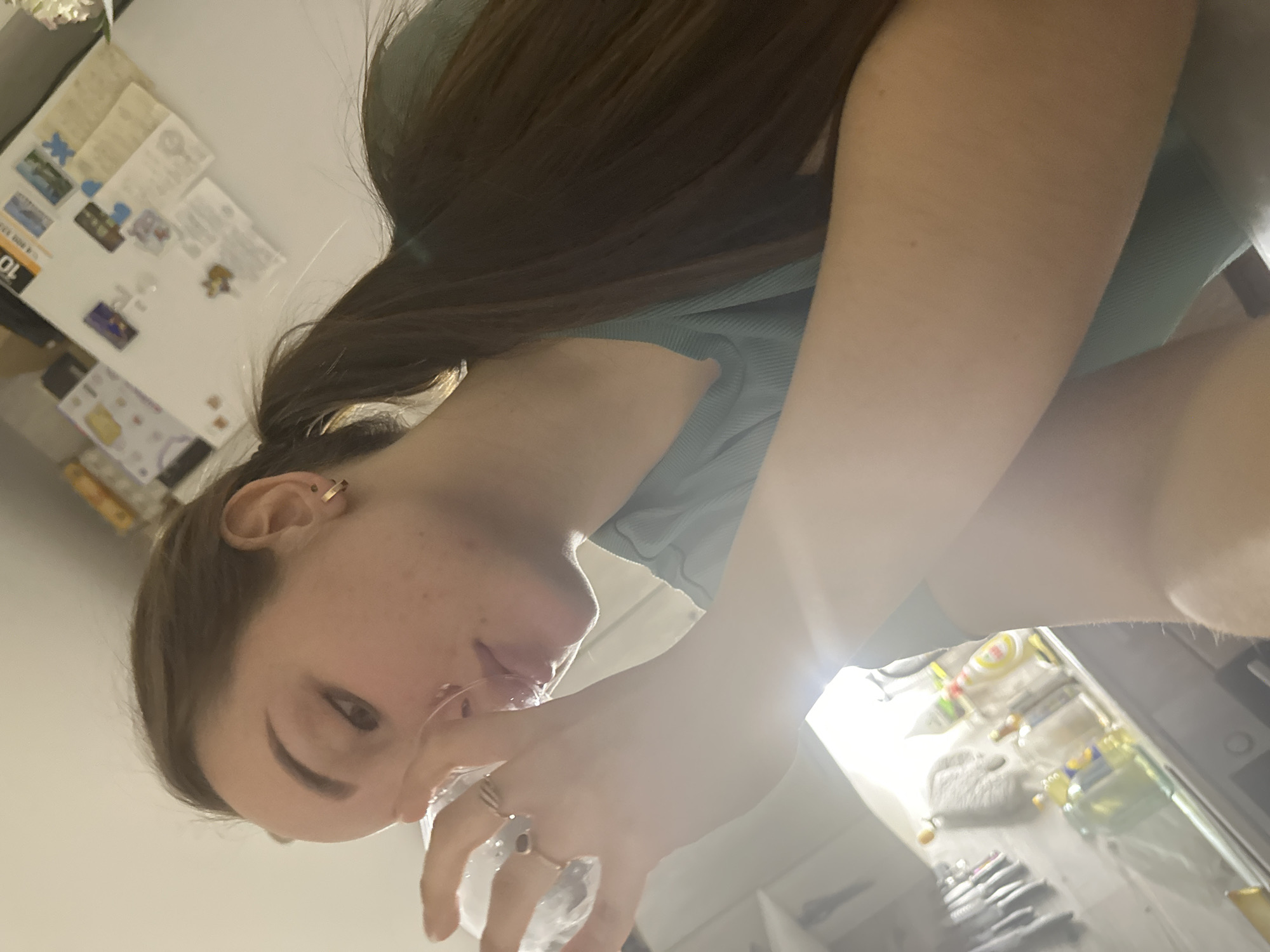 A woman with brown hair in a ponytail and wearing a light blue ribbed top is holding a clear glass of water in her right hand, drinking from it. She has a gold hoop earring in her left ear and two rings on her right hand. Behind her is a white refrigerator door with various magnets and papers attached. The kitchen contains stainless steel cookware and various bottles and containers. The location is not evident to be in the city of Chaikovsky, Russia.