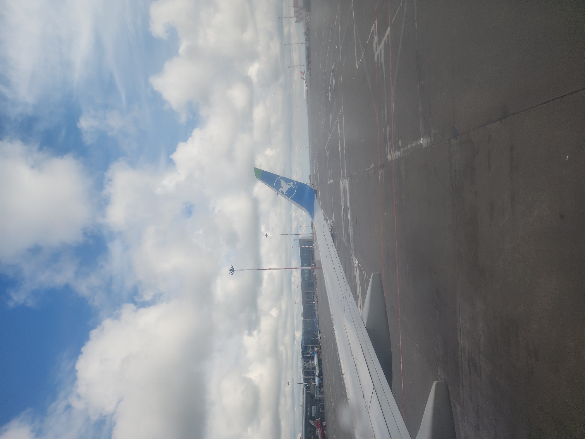 The image shows a view from an airplane window, likely at an airport in Bol'shie Kabany, Russia. The left side of the frame is dominated by a bright blue sky with scattered white clouds. In the lower part of this section, the blue tail fin of the airplane is visible, emblazoned with a light-colored emblem. The right side of the image displays a section of the airplane wing, painted white, with visible flaps and components. Beyond the wing, the airport tarmac stretches into the distance, appearing as a dark gray surface with faint lines. Various airport structures, including buildings and light poles, are visible along the horizon line. The overall scene conveys the feeling of preparing for takeoff or shortly after landing.