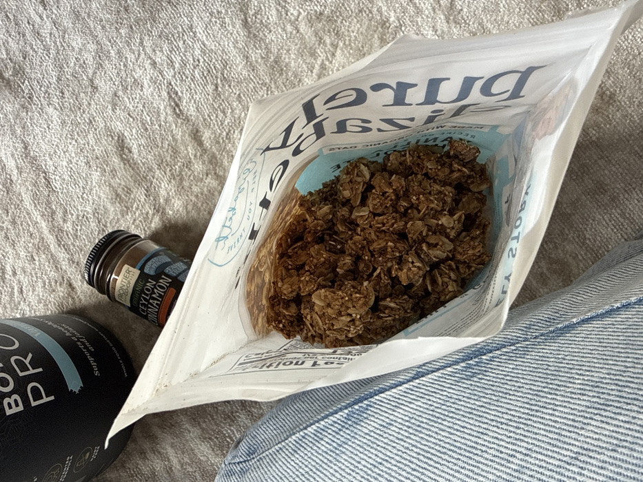 The image presents an overhead perspective of several items positioned on a light-colored, textured fabric surface. A white, open stand-up pouch, containing dark brown granola or a similar clustered food product, dominates the central foreground. Text visible on the pouch includes a partial 'HERITAGE' in blue and nutritional information on the opposing side. To the upper left, a black cylindrical container, partially obscured, displays 'BONE PRO' and 'Supports a healthy bone and joint.' A small brown glass jar with a black lid, labeled 'FRONTIER CO-OP ORGANIC CEYLON CINNAMON,' rests between the supplement container and the granola pouch. A segment of a human leg, clad in light blue denim jeans, is visible along the left frame edge. This scene is located in the city of Dubai, United Arab Emirates.