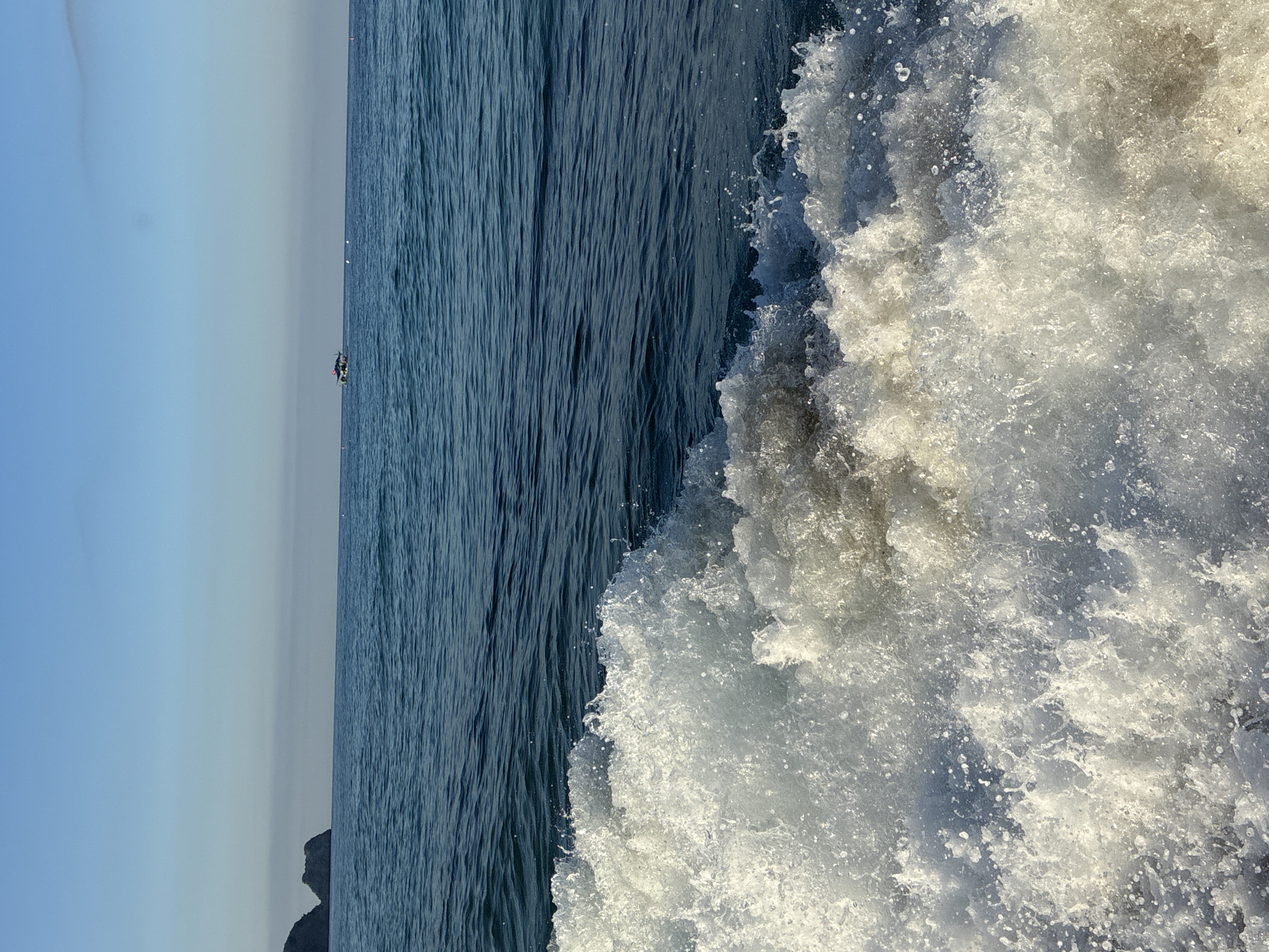 The image captures a dynamic ocean scene in Alanya, Turkey, with a focus on a crashing wave in the foreground. The wave is a cascade of white foam and spray, obscuring the water beneath. In the mid-ground, the deep blue sea stretches towards the horizon, with gentle ripples suggesting a calm day. A small boat with a dark hull and a lighter top is visible in the distance, navigating the open water. On the far left of the horizon, a rocky outcrop, characteristic of Alanya's coastline, juts out from the sea, partially shrouded in a light haze. The sky above is a clear, pale blue, with a subtle gradient towards a lighter hue near the horizon. No people are visible in the image.