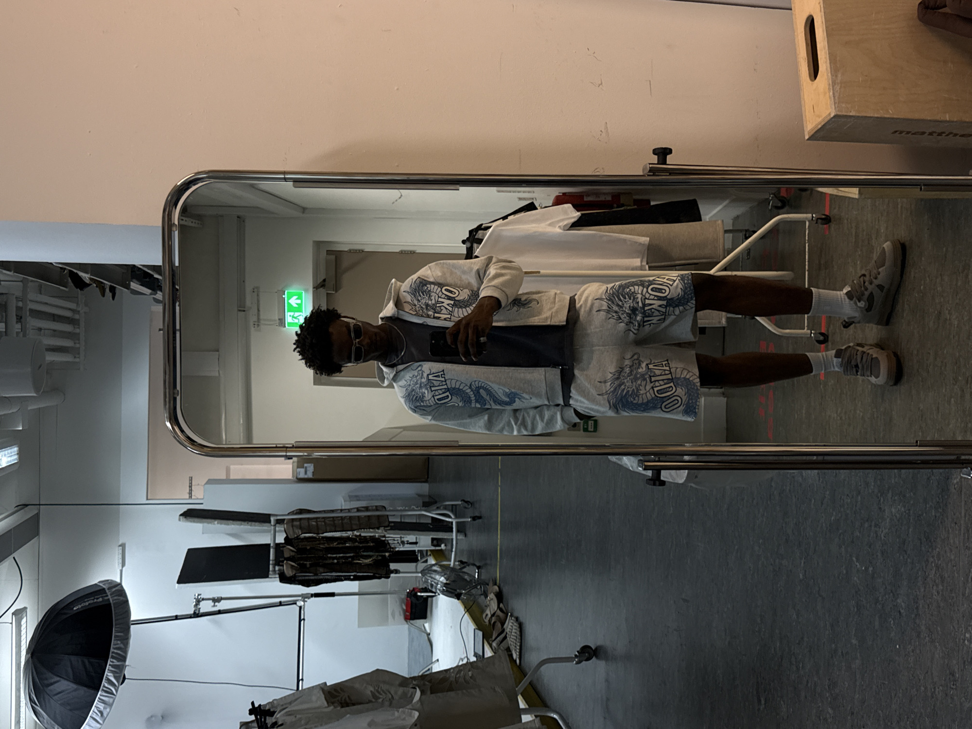 The image depicts a man reflecting in a large, free-standing mirror. The man is wearing a matching grey hoodie and shorts with a dragon design, a dark grey undershirt, white socks, and grey sneakers. He is holding a smartphone, presumably taking the photo. The mirror is set in what appears to be a studio or storage space with various equipment, including lighting umbrellas, garment racks with clothes, and a wooden box labeled "matthews". A green emergency exit sign is visible in the background. Based on the presence of the "matthews" box, which is a popular brand in the film and photography industry, the location is likely a studio space used for photoshoots or film production, but the presence of garment racks also suggest a retail outlet that uses the studio to take advertising images. The location is not specifically identifiable as being in London. The scene is brightly lit.