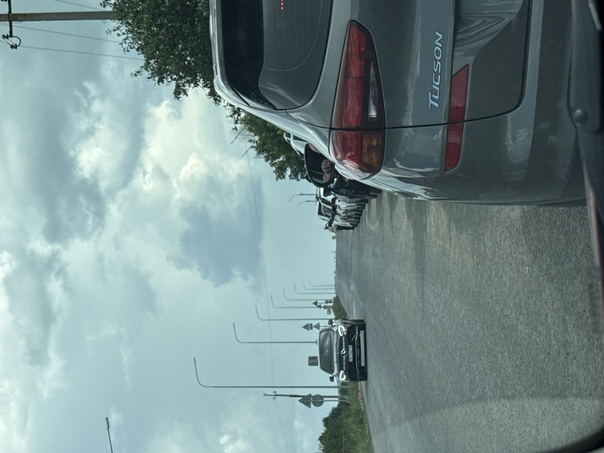 The image depicts a traffic jam on a road in Kachimir, Russia, under a cloudy sky. A line of cars is stationary on the asphalt road, with trees and utility poles lining the sides. In the foreground, the rear of a grey SUV is visible, with the "TUCSON" logo clearly displayed on the tailgate. Further down the road, a black Mercedes-Benz is approaching the traffic. Several streetlights are also visible along the road, indicating it's likely a main thoroughfare. The overall scene suggests a typical day in a city where traffic congestion is common.