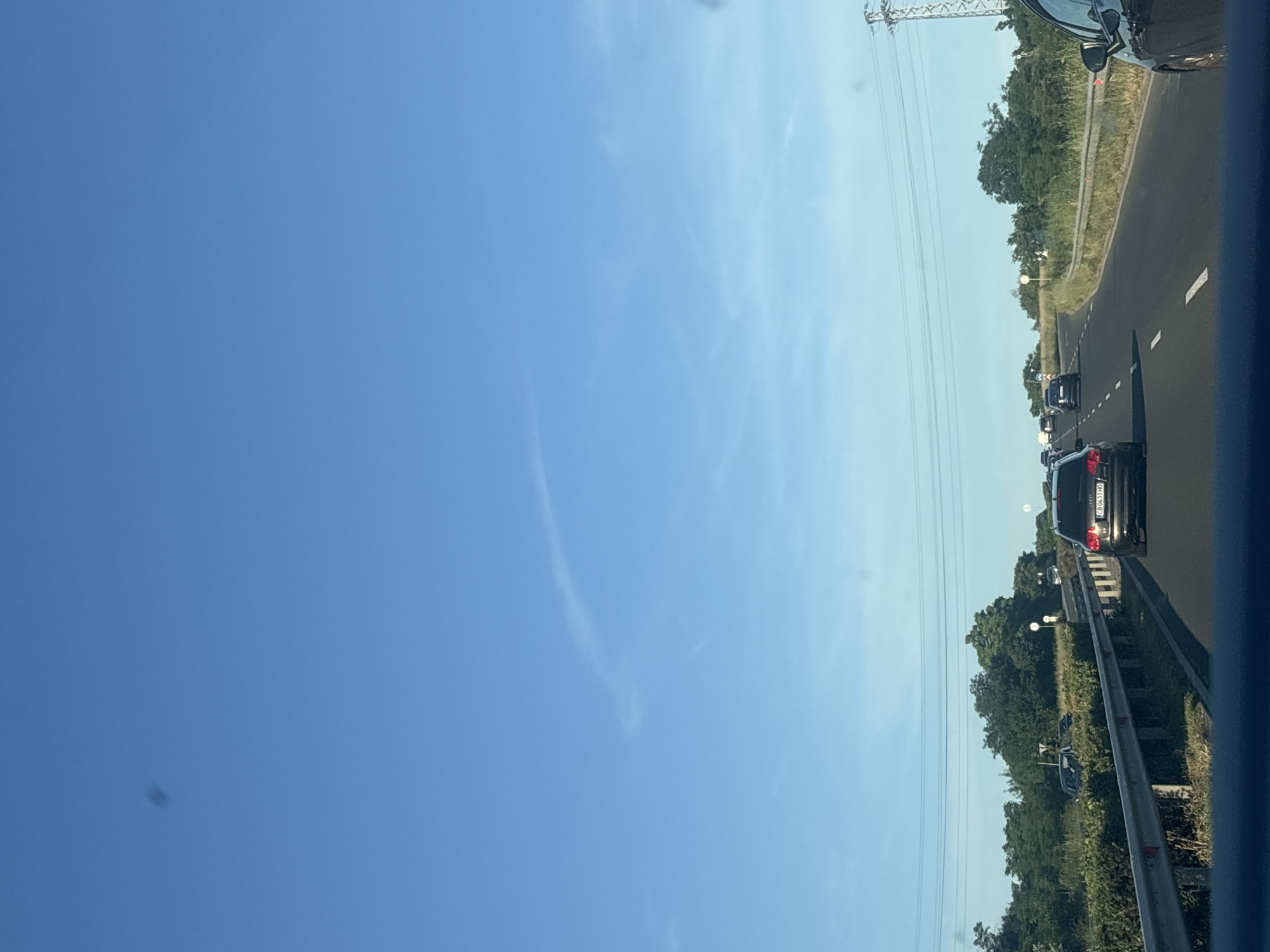 The image depicts a traffic jam on a highway, likely in Burgas, Bulgaria. The perspective is from inside a vehicle, looking forward. The scene is dominated by a clear blue sky with faint wispy clouds. The highway is visible, with several cars in front, indicating slow-moving traffic. The road has white lane markings and a barrier on the right side.
Green trees and vegetation line the sides of the highway, with a tall electrical pylon visible in the distance. The sun appears to be high in the sky, suggesting it is daytime, likely late morning or afternoon, given the bright conditions and clear visibility. There are no people clearly visible within the frame, but the presence of multiple vehicles implies numerous occupants.
A notable detail on the license plate of the rearmost car reads "CB 631 HO". The overall mood is one of a mundane, everyday occurrence - being stuck in traffic on a sunny day. The image does not convey any strong emotions, other than perhaps a sense of mild frustration or patience associated with traffic delays. The setting is clearly a road, and the presence of the highway infrastructure points to a developed area.