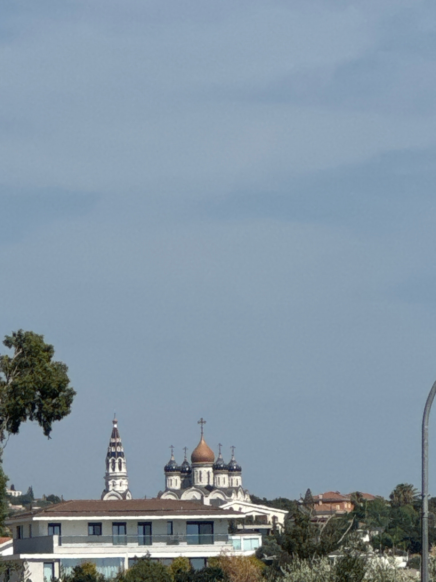 The image shows a skyline with multiple buildings and a clear sky. In the foreground, a modern white building with a flat roof and large windows is visible. Behind this building, a traditional church complex with multiple domes and a tall bell tower stands out. The church features onion domes, some of which are copper-colored, and a prominent cross on top. The bell tower is white with black accents and a pointed spire. To the left of the church complex, a large tree with green foliage partially obscures the view. The background reveals a hilly landscape with more buildings and scattered trees. A street light pole is visible on the far right of the image. The architecture of the church suggests it might be an Eastern Orthodox church. This scene is likely located in the city of Germasogeia Municipality, Cyprus, known for its diverse architectural styles and proximity to religious sites. The lighting suggests it is daytime, with the sun casting soft shadows. There are no people visible in the image.