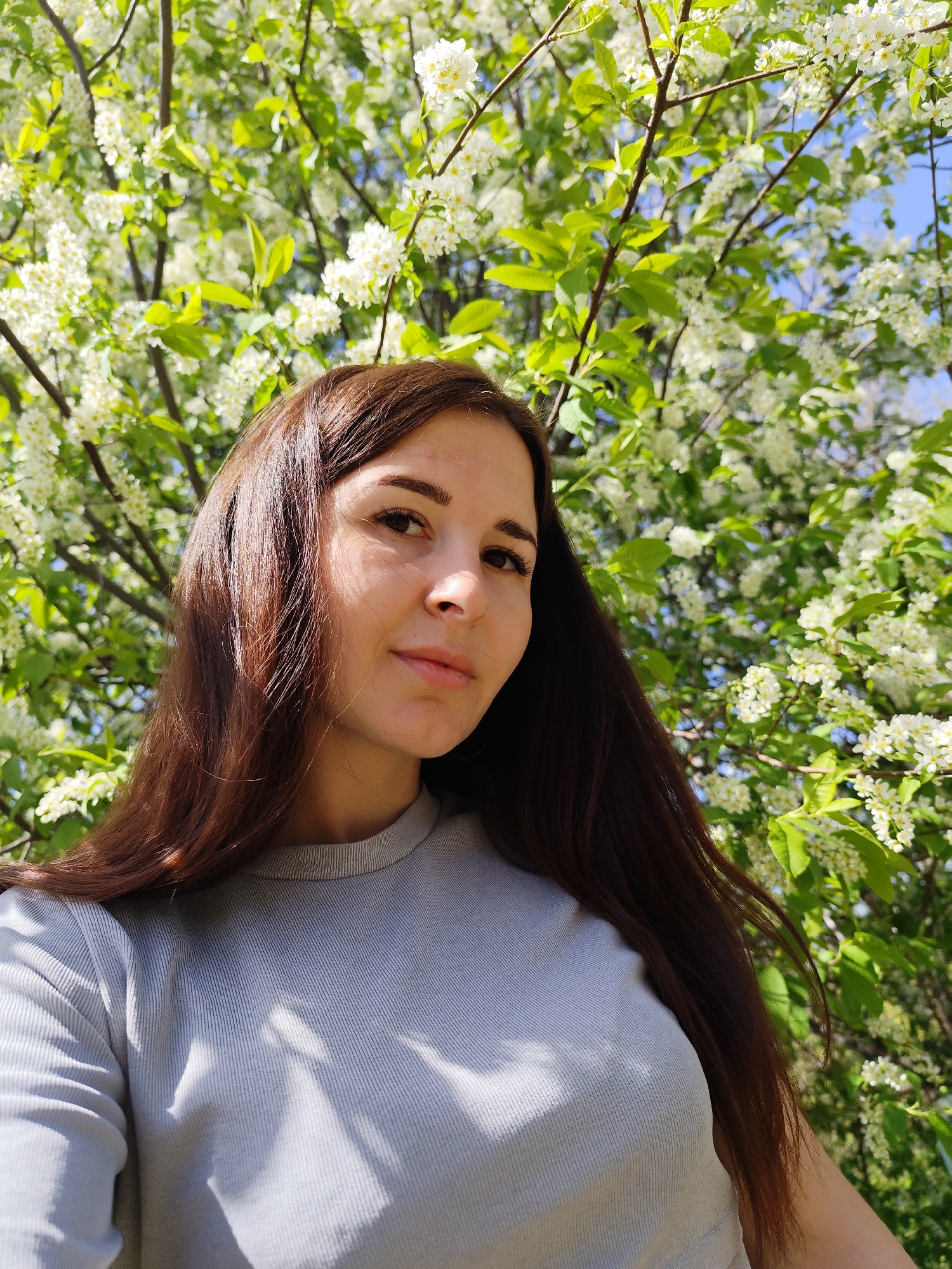 A woman with long, brown hair is standing in front of a tree with white blossoms. She is wearing a gray long-sleeved shirt. The location is likely outdoors, with the tree and blossoms suggesting a park or garden. There is no indication of the location being in the city of Фоки, Russia. The woman is looking at the camera.