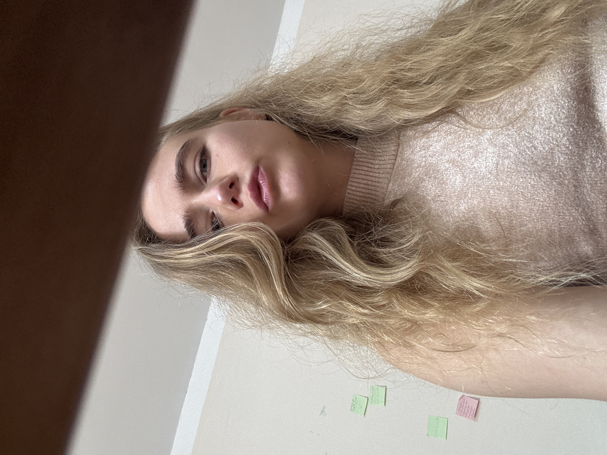 A low-angle shot features a woman with blonde hair and light complexion. She's wearing a beige sweater. Her face is centered in the frame, with her gaze directed slightly downward. A dark, out-of-focus object occupies the upper left corner. The background is a plain white wall with a few small, square sticky notes attached to it. The city and specific location are unknown.