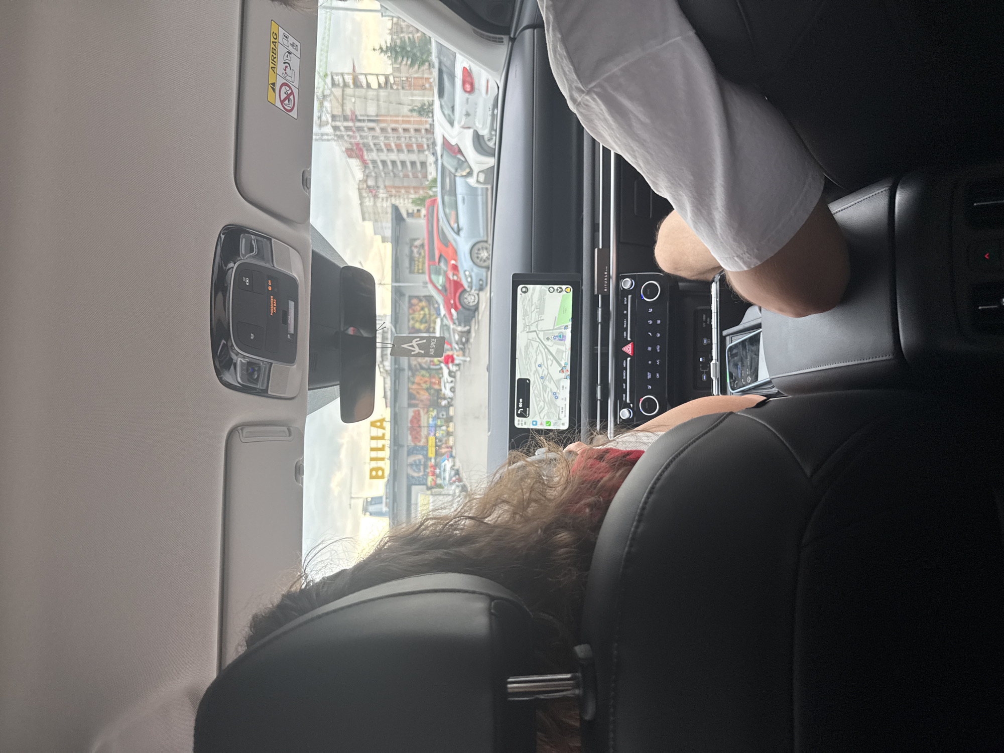 The image captures the interior of a vehicle from the back seat, looking forward through the windshield at an outdoor scene in Sofia, Bulgaria.
In the foreground, the backs of two front seats are visible. The left seat is occupied by a person with long, curly, dark brown hair, appearing to wear a red garment. The right seat is occupied by a person with fair skin, wearing a white short-sleeved shirt and shorts, with their right arm resting on the center console. The car's interior features a gray headliner, overhead console with "PASSENGER AIR BAG ON" indicator, and a sun visor with a prominent "AIRBAG" warning label. An "AIR SPICE" car air freshener hangs from the rearview mirror. The dashboard is modern, featuring a wide, integrated infotainment screen displaying a map or navigation system. Below the screen are touch-sensitive climate controls and physical buttons, with "RITUALS..." branding visible on the trim.
Through the windshield, a large supermarket building with the prominent yellow "BILLA" signage is visible. Below the "BILLA" sign, the building's facade features colorful murals or graffiti. A parking lot in front of the BILLA store contains several parked vehicles, including a red van, a light blue hatchback, and a white SUV. In the mid-ground and background to the right, a significant urban development is underway, characterized by multiple multi-story buildings under construction with scaffolding and a tall green construction crane. The sky is partly cloudy.
The location is identified as a **BILLA supermarket on Tsarigradsko Shose Boulevard in Sofia, Bulgaria**, situated in an area experiencing substantial new construction.