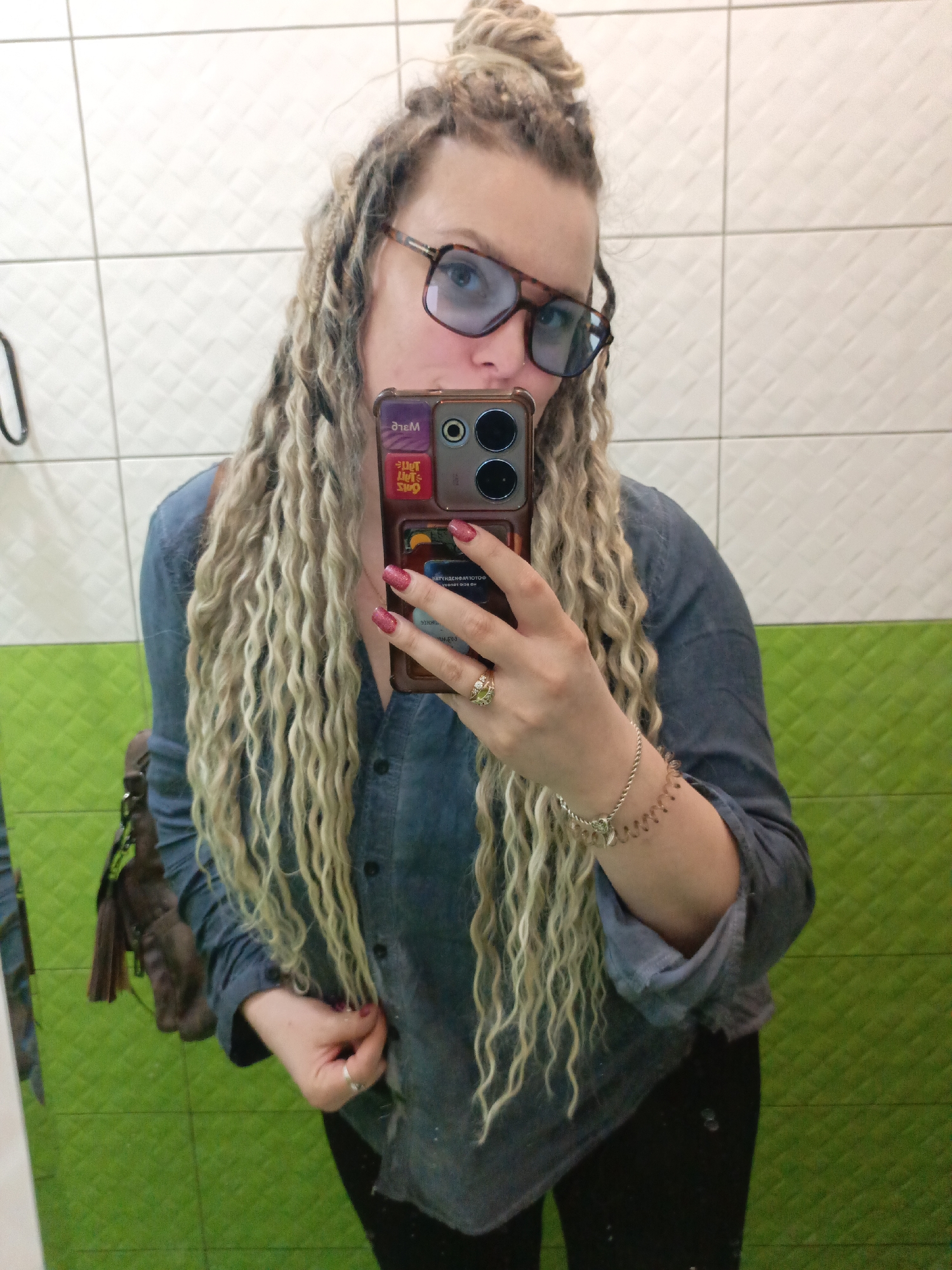 The image depicts a woman taking a selfie in a bathroom. She has light-blonde dreadlocks piled up in a bun on top of her head, and wears blue-tinted glasses. She is wearing a denim shirt and has a small brown bag slung over her shoulder. She holds a phone in her hand, with a phone case adorned with stickers. Her nails are painted with glittery red polish, and she is wearing a ring and bracelet. The bathroom has white and green tiled walls. The location is not specified as Verkh-Sosnova, Russia, in the image.