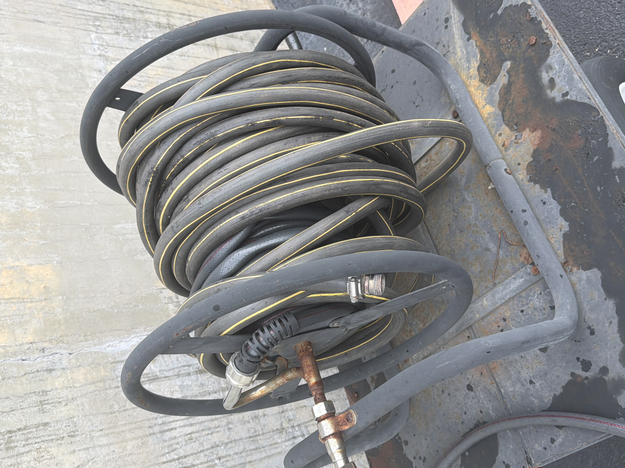 An outdoor, medium-shot, eye-level photograph shows a heavy-duty, black rubber hose coiled onto a metal hose reel. The hose has two parallel yellow stripes running along its length. The reel is mounted on a wheeled cart, also made of dark metal. The cart appears to be positioned outdoors, on a grey, weathered surface with visible rust spots and water stains. In the background, a rough, textured concrete wall is visible. A rusted metal coupling is attached to the end of the hose, with another silver metal fitting connected to it. The overall scene suggests an industrial or maintenance setting in Murfreesboro, United States of America.