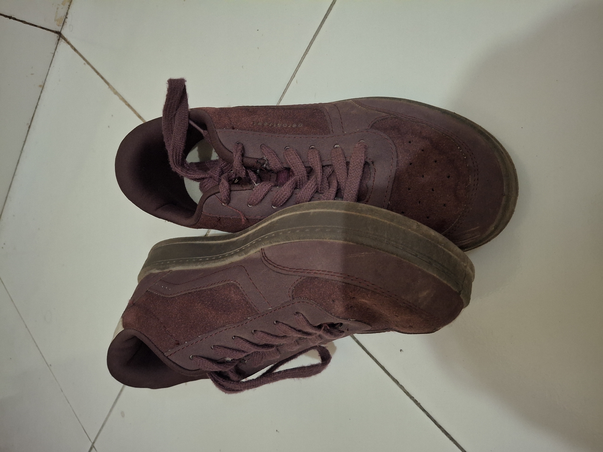 The image shows a pair of maroon low-top sneakers placed on a white tiled floor. The shoes are angled, with the left shoe in the foreground and the right shoe behind it and slightly to the right. The shoes appear to be made of a suede-like material and have white soles. The laces are a darker shade of maroon. The floor tiles are square and arranged in a grid pattern. There are faint shadows cast by the shoes. The background is out of focus. The scene is indoors. The image was taken in Banguntapan, Indonesia.