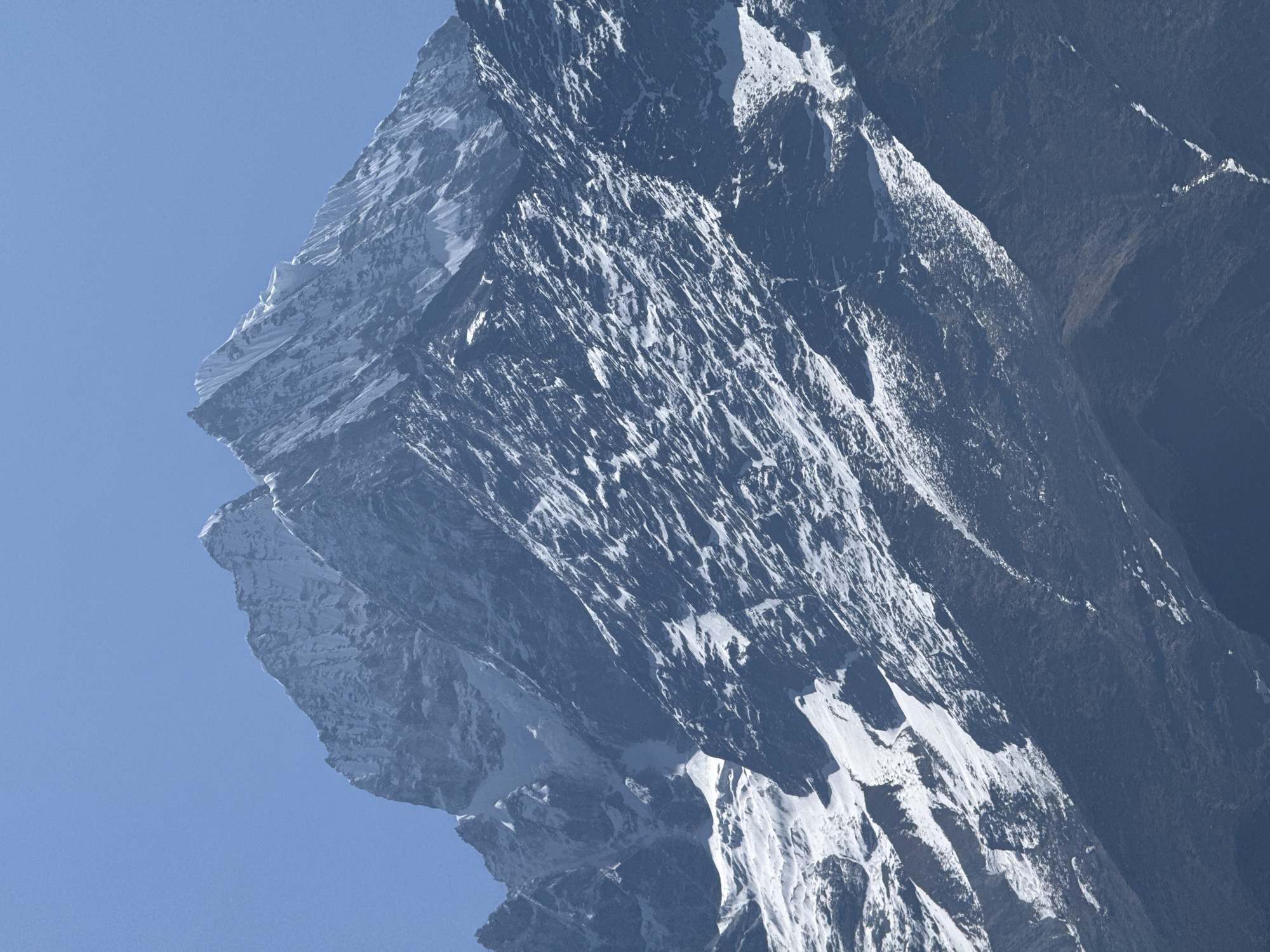The image presents a close-up view of a mountain face, likely located near Namche, Nepal, given the context. The mountain is characterized by steep, rocky terrain with extensive snow and ice cover, particularly on its upper and more exposed slopes. Sharp ridges and outcroppings define its contours. Patches of bare rock and shadow are visible in the lower section, suggesting areas where the snow has melted or where the terrain is too steep for accumulation. The sky above is a clear, light blue, providing a stark contrast to the darker tones of the mountain. There are no identifiable people, objects, actions or interactions visible in the scene.