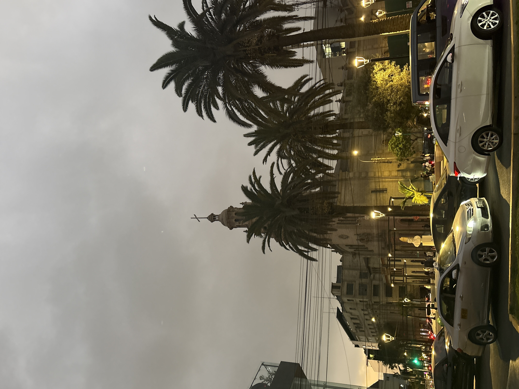 The image depicts a street scene in San Isidro, Peru, during twilight. Tall palm trees are prominent in the foreground, with their fronds casting shadows. In the background, a church with a cross-topped steeple is visible, along with several multi-story buildings. The street is populated with various vehicles, including sedans, a van, and what appears to be a taxi. Pedestrians are sparsely scattered on the sidewalks and near some of the buildings. Streetlights illuminate the scene, casting a warm glow on the surroundings. The sky is overcast with a greyish hue, suggesting a cloudy evening. The overall atmosphere is that of a bustling urban environment transitioning into night.