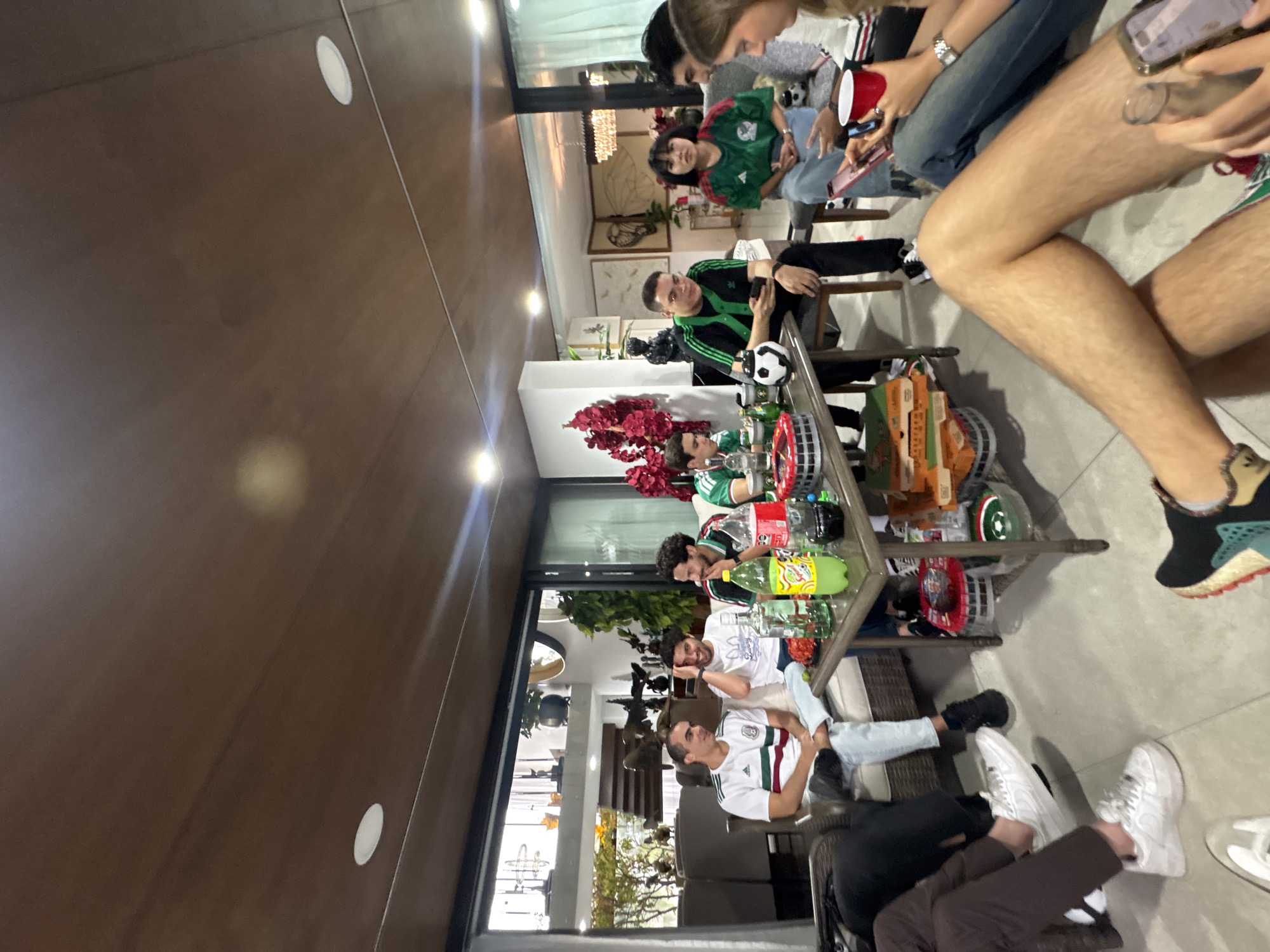 A group of young adults are gathered in a modern living room, likely in the city of San Bernardino Tlaxcalancingo, Mexico, to watch a sporting event. The room features a wooden ceiling with recessed lighting, large glass doors opening to an outdoor area, and contemporary furniture. The individuals, dressed in casual attire, with several sporting soccer jerseys, are seated on a couch and chairs around a coffee table laden with food and drinks, including pizza boxes, bottles of soda, and alcoholic beverages. Some are actively engaged in conversation, while others are looking at their phones, suggesting a relaxed and social atmosphere. A soccer ball is visible on the coffee table, reinforcing the sports theme. The overall impression is one of a casual gathering among friends, possibly celebrating a victory or enjoying a game together.