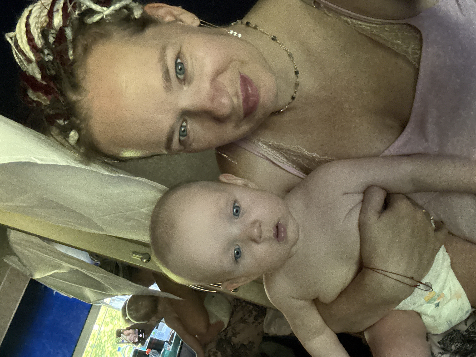 A woman with dreadlocks and blue eyes, wearing a pink top and a beaded necklace, holds a baby with blue eyes. The baby is shirtless and wearing a diaper. In the background, another baby is visible. A smartphone is being held up to take a photo, showing a screen with an image of the two babies. The setting appears to be indoors, possibly a home, with a view of greenery through a window. The image is a selfie taken by the woman. The location is specified as Gal'chino, Russia.