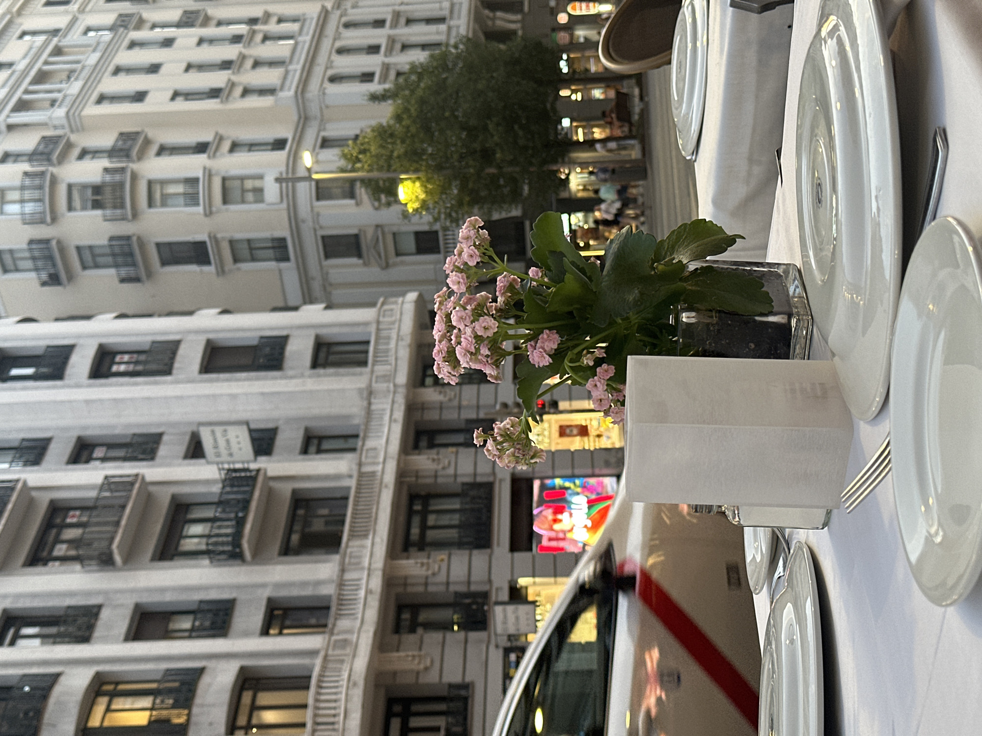 The scene depicts an outdoor restaurant setting in Madrid, Spain, with a focus on a dining table. In the foreground, there is a set table with white plates, cutlery, and a glass vase containing pink flowers and green leaves. A white napkin is placed near the vase. The restaurant seems to be on a street with buildings on one side. The buildings are multi-storied with many windows and balconies. One building has a fire escape. In the background, there's a street with people and businesses. A car is partially visible on the right side of the image, with a reflection of a person in the side window. The overall ambiance suggests a lively urban setting.