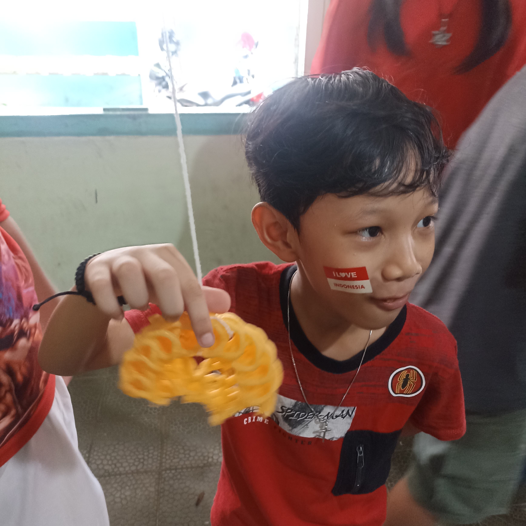 In Magelang, Indonesia, a young boy with dark hair is shown in a close-up shot. He is wearing a red t-shirt with a black collar and a silver necklace. A sticker with the Indonesian flag colors and the words "I LOVE INDONESIA" is placed on his cheek. In his right hand, he holds a yellow, circular snack with a textured surface. The background is slightly blurred, showing a wall with a textured pattern and a hint of a window with blue and white light. Another person, wearing a red shirt, is partially visible behind him, and another person is to the right, wearing a gray shirt.