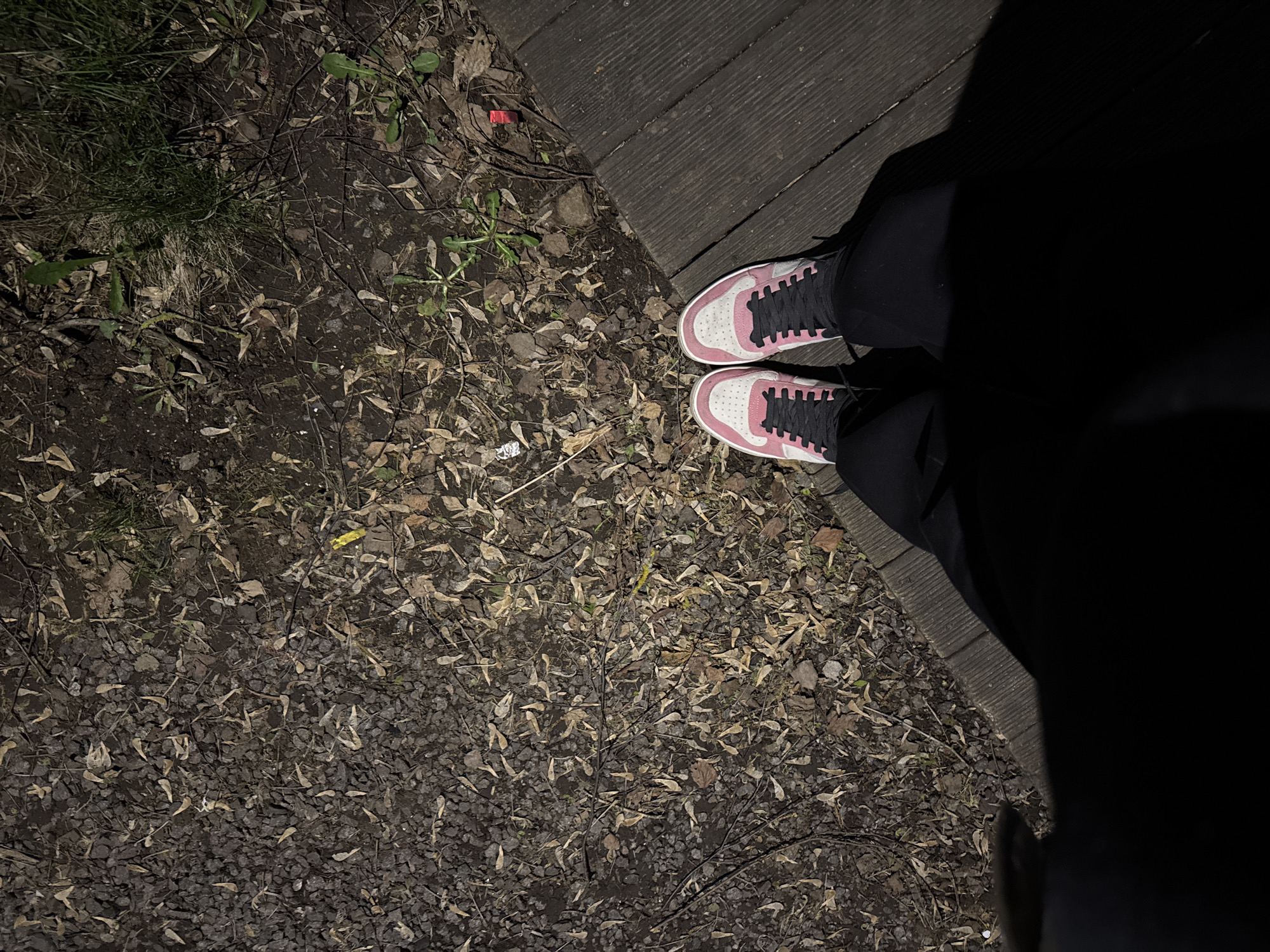 An overhead, low-light shot captures a person's feet and the ground below them. The person is wearing black pants and pink and white sneakers with prominent black laces, standing on what appears to be a gravel path or natural ground.
The ground surface is a mix of small dark gravel, dried leaves (many resembling maple samaras), small twigs, and patches of sparse green grass and weeds. To the right, a dark wooden surface, possibly decking or a wooden path, meets the natural ground at an angle. A significant portion of the image, particularly to the right and bottom, is covered by a deep, dark shadow, suggesting a low light environment, possibly evening or night, with an unseen light source illuminating the visible areas.
Scattered across the ground are a few small pieces of debris: a red rectangular object near the wooden planks, a small yellow item further to the left, and a tiny white piece near the left shoe. There is no discernible activity other than the person standing. No text is visible in the image.