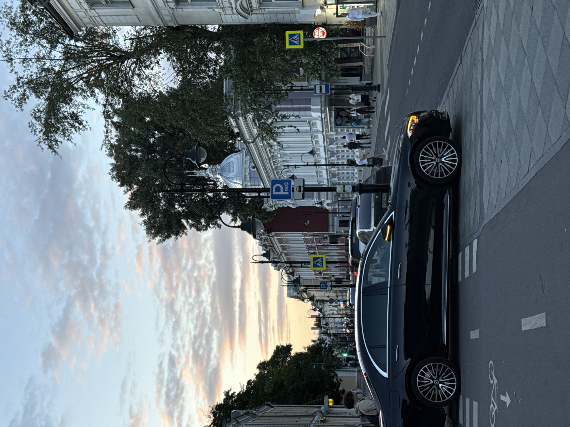 The image captures a street scene in Moscow, Russia, during dusk. The sky is filled with soft, pastel clouds, indicating the sunset. A black Mercedes-Benz sedan is prominently positioned in the foreground, partially obscuring a pedestrian crossing marked with white stripes and a bicycle lane symbol.
The street is lined with buildings of varying architectural styles. On the left, older, muted-colored buildings with classic facades are visible. On the right, a more ornate building with a light purple facade, white trim, and decorative columns stands out, featuring a distinctive silver dome. Streetlights with antique designs line the street, casting a warm glow.
Several people are present on the sidewalk. Some are walking, while others appear to be standing and conversing. A man in a white suit is visible near the right edge of the frame, standing by a lamppost. Traffic is present on the street, with other cars visible in the distance, suggesting a moderately busy urban environment. Road signs, including parking signs and pedestrian crossing signs, are scattered along the street. The overall impression is of an urban street transitioning from day to night, with a mix of historical and modern elements.