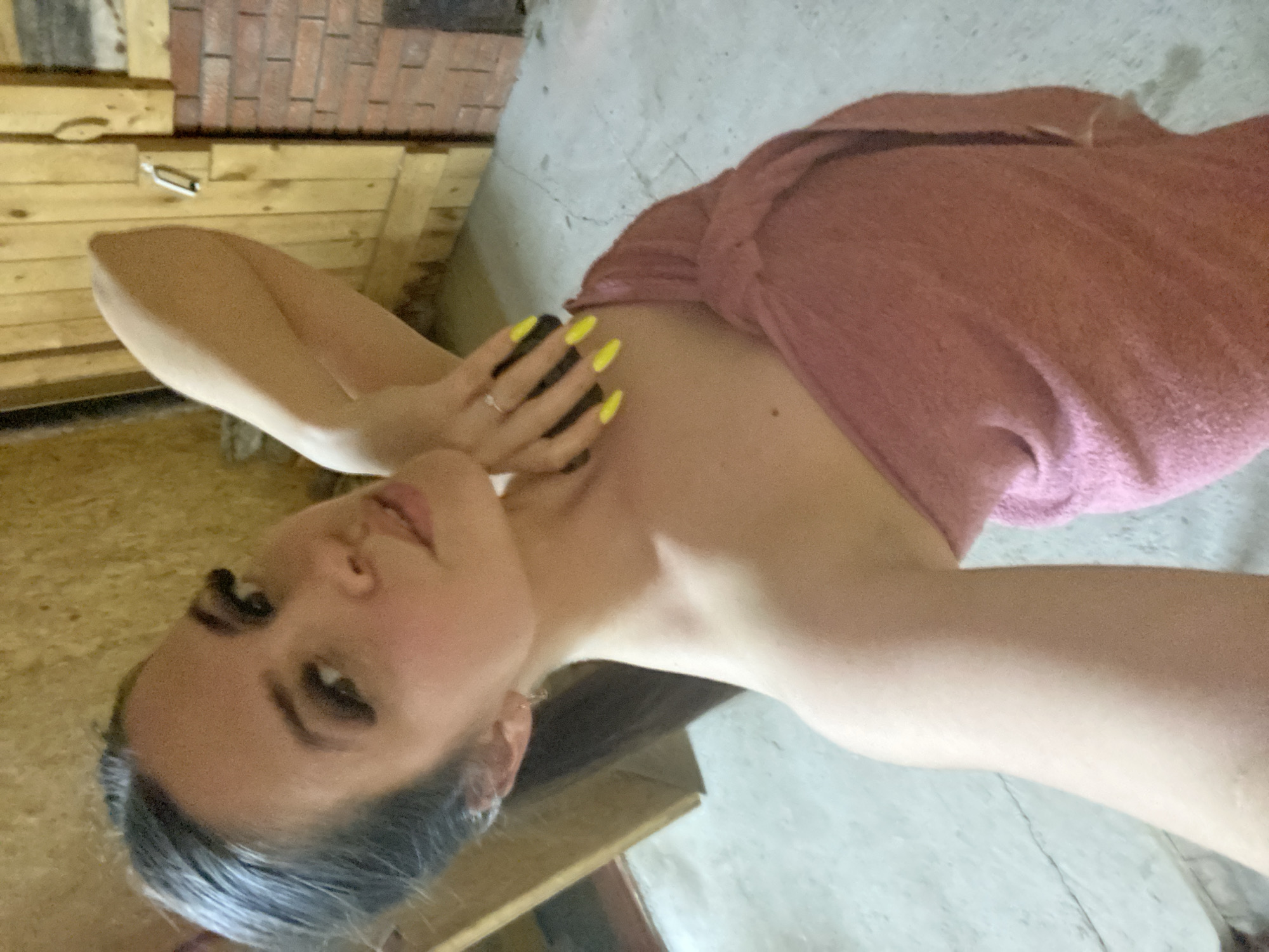 A woman with gray hair, wearing heavy dark eye makeup, is seen from the shoulders up. She is wrapped in a pink towel. Her left hand is raised near her face, displaying a ring and yellow nail polish. The background consists of wooden planks and a gray concrete surface. The location and city are not discernible from the image.