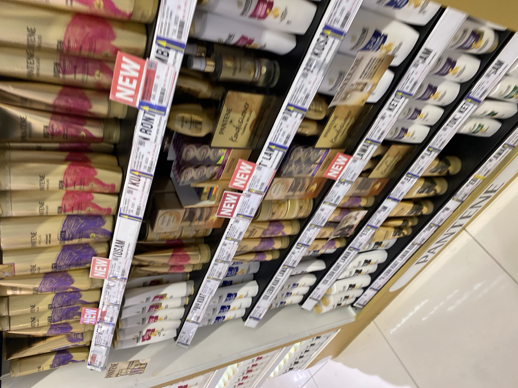 The image shows a retail display shelf stocked with Pantene hair care products in a store. The products are arranged in rows on multiple shelves. Various Pantene bottles, tubes, and boxes are visible, many featuring gold and purple packaging. Some shelves have price tags or promotional labels, with "NEW" tags prominently displayed on some items. The environment appears to be indoors, likely a supermarket or pharmacy, with a tiled floor visible at the bottom of the image. The lighting is bright and diffused, typical of a retail setting. There are no people or interactions depicted in the image, focusing solely on the product display. The visible text on some labels suggests the location is in Banda Aceh, Indonesia, with phrases like "TAK LEBIH KUAT LAWAN" and "SAM" visible.