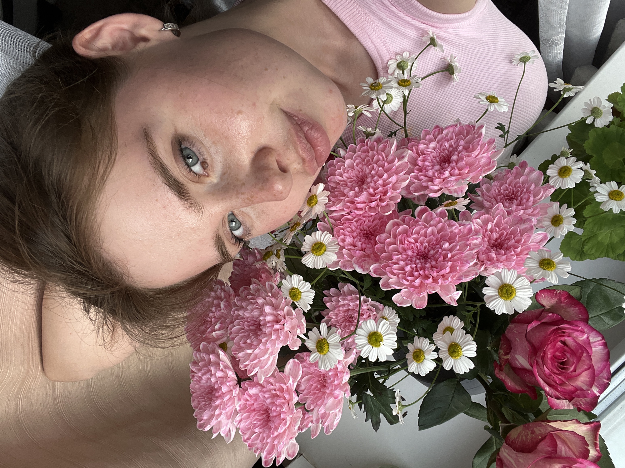 A young Caucasian woman with brown hair and blue eyes is lying down with a bouquet of pink chrysanthemums, white daisies, and pink roses. She is wearing a pink ribbed tank top and has a silver hoop earring in her right ear. The background appears to be a textured, neutral-colored wall. The scene is indoors, near a window, and a white surface is visible in the lower portion of the image. There is no indication of the specific location in Chaikovsky, Russia.
