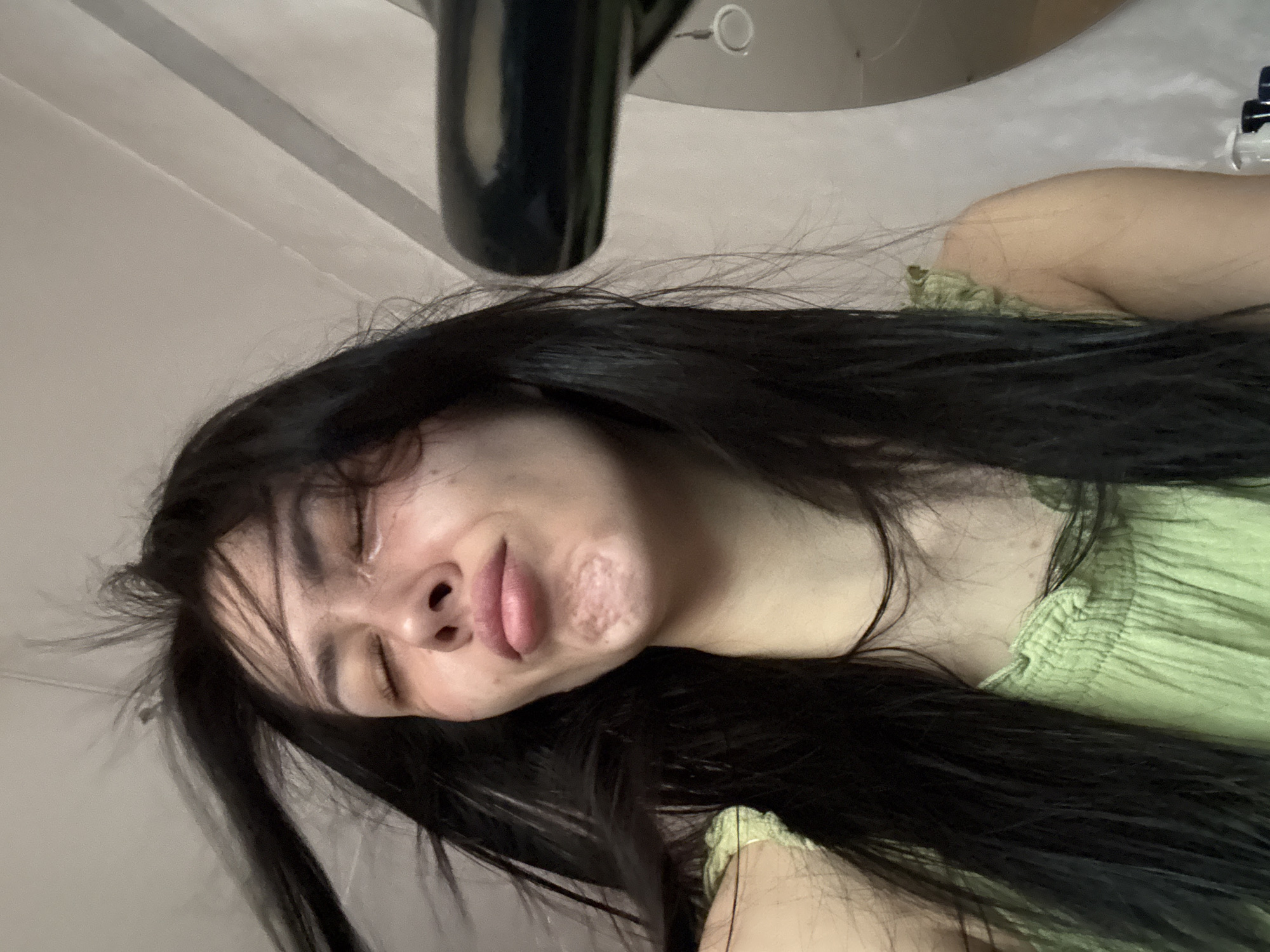 A close-up, angled shot captures a woman with long, black hair lying down. Her eyes are closed and her face is turned slightly towards the camera, revealing some skin texture and a mark on her cheek. She is wearing a light green garment with ruffled detailing. Above her head is a black object, possibly a part of a fixture or appliance. The background appears to be a plain wall. There's no information to discern its location is in the city of Votkinsk, Russia.