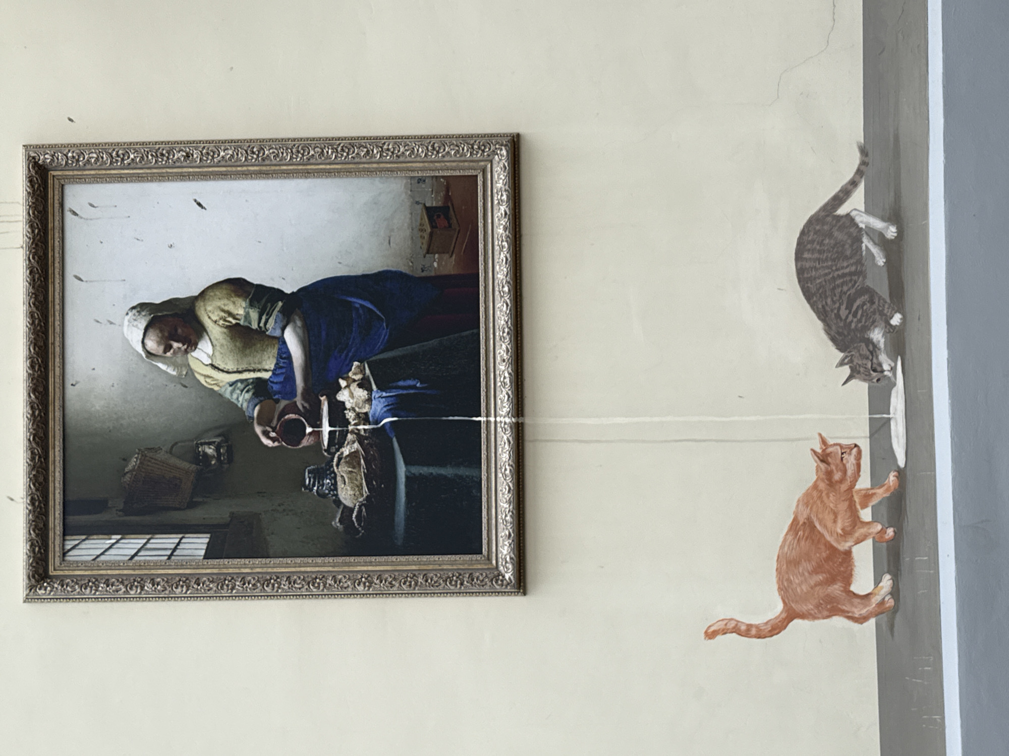 A wall in a building in Moscow, Russia, features a framed reproduction of Johannes Vermeer's "The Milkmaid" alongside a painted mural of two cats interacting with a white disc resembling a saucer. A string appears to extend from the painting to the mural, visually connecting the woman in the artwork to the cats. The wall has a cream color and a gray stripe.