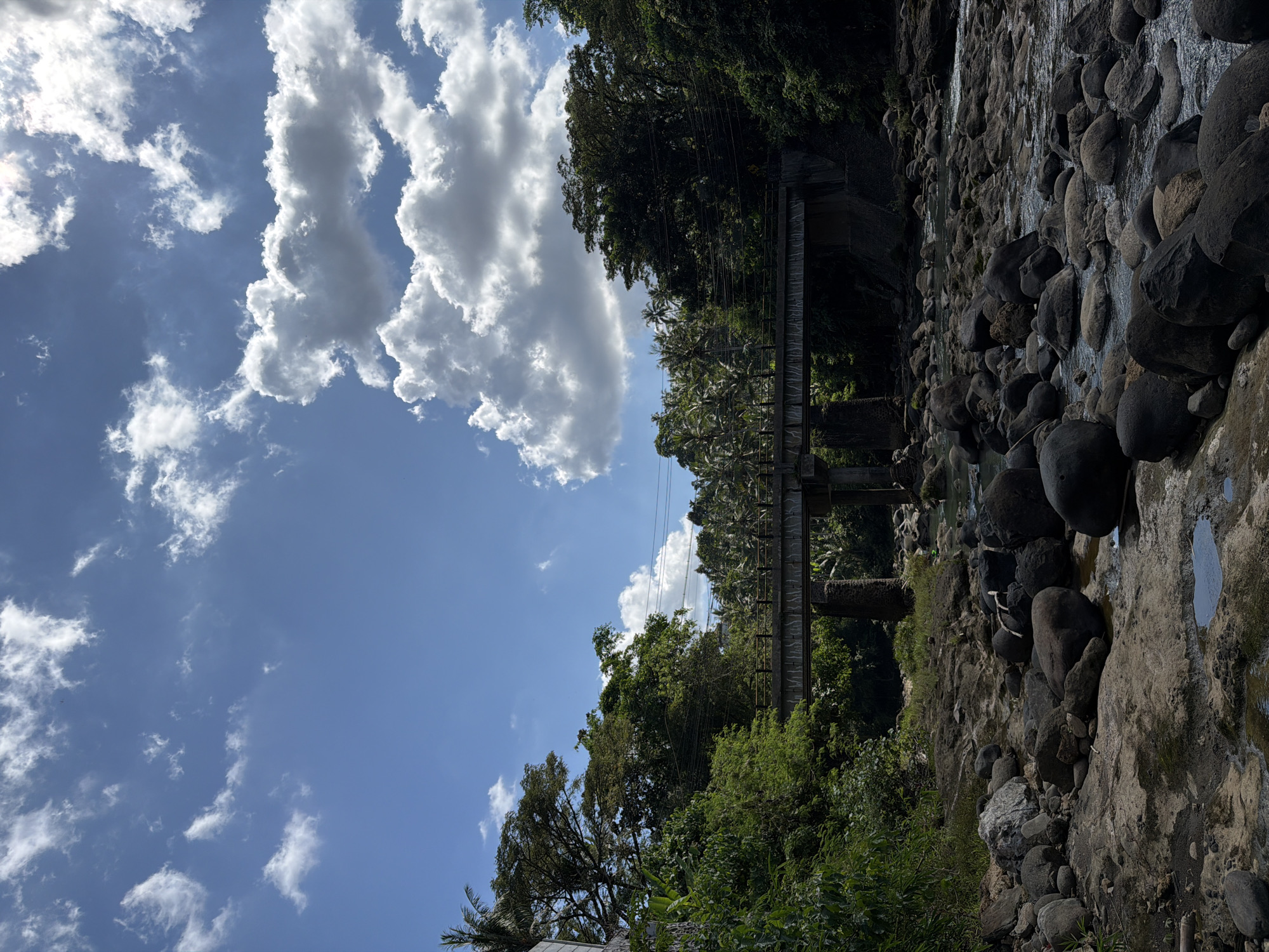 The image presents a view in Selemadeg Timur, Indonesia. The sky is blue with cumulus clouds, occupying the left side of the frame. On the right side, a rock formation descends from top to bottom. A pedestrian bridge crosses the top of the rock formation, shrouded in green vegetation. The environment is lush with tropical trees. A river runs parallel to the rock formation.