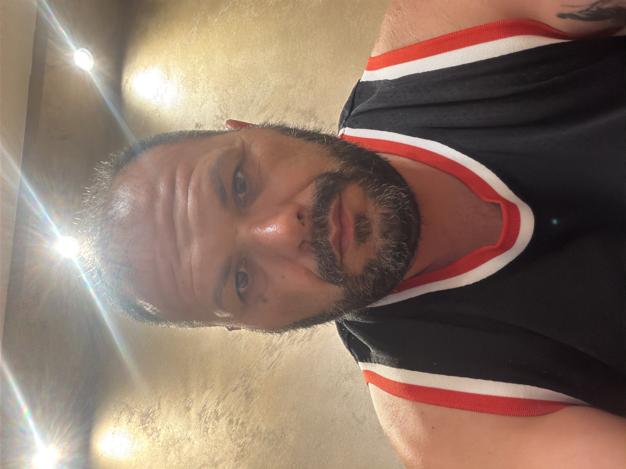This is a selfie of a man with short, dark hair and a beard, wearing a black sleeveless jersey with red and white trim. He has fair skin and is looking directly at the camera with a neutral expression. The background appears to be an indoor setting with a textured, light-colored wall and bright overhead lights causing some glare. There is no discernible information about the location in Burgas, Bulgaria.