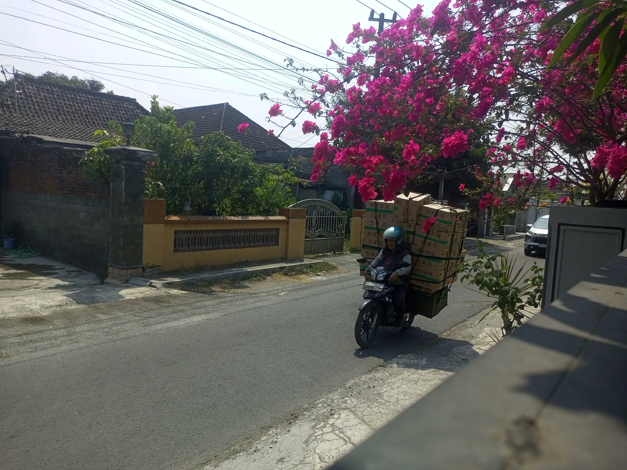 A person is riding a motorcycle on a paved road in the city of Ngasem, Indonesia. The motorcycle is carrying a large stack of boxes secured to the back. The person is wearing a blue helmet and appears to be in motion. The surroundings include houses with tiled roofs, a brick wall, a metal gate, and a tree with bright pink flowers. Overhead, a tangle of electrical wires is visible against a clear sky. A white car is parked on the side of the road in the background. The road surface is asphalt with some lighter colored patches and debris. The lighting suggests it is daytime with bright sunlight.