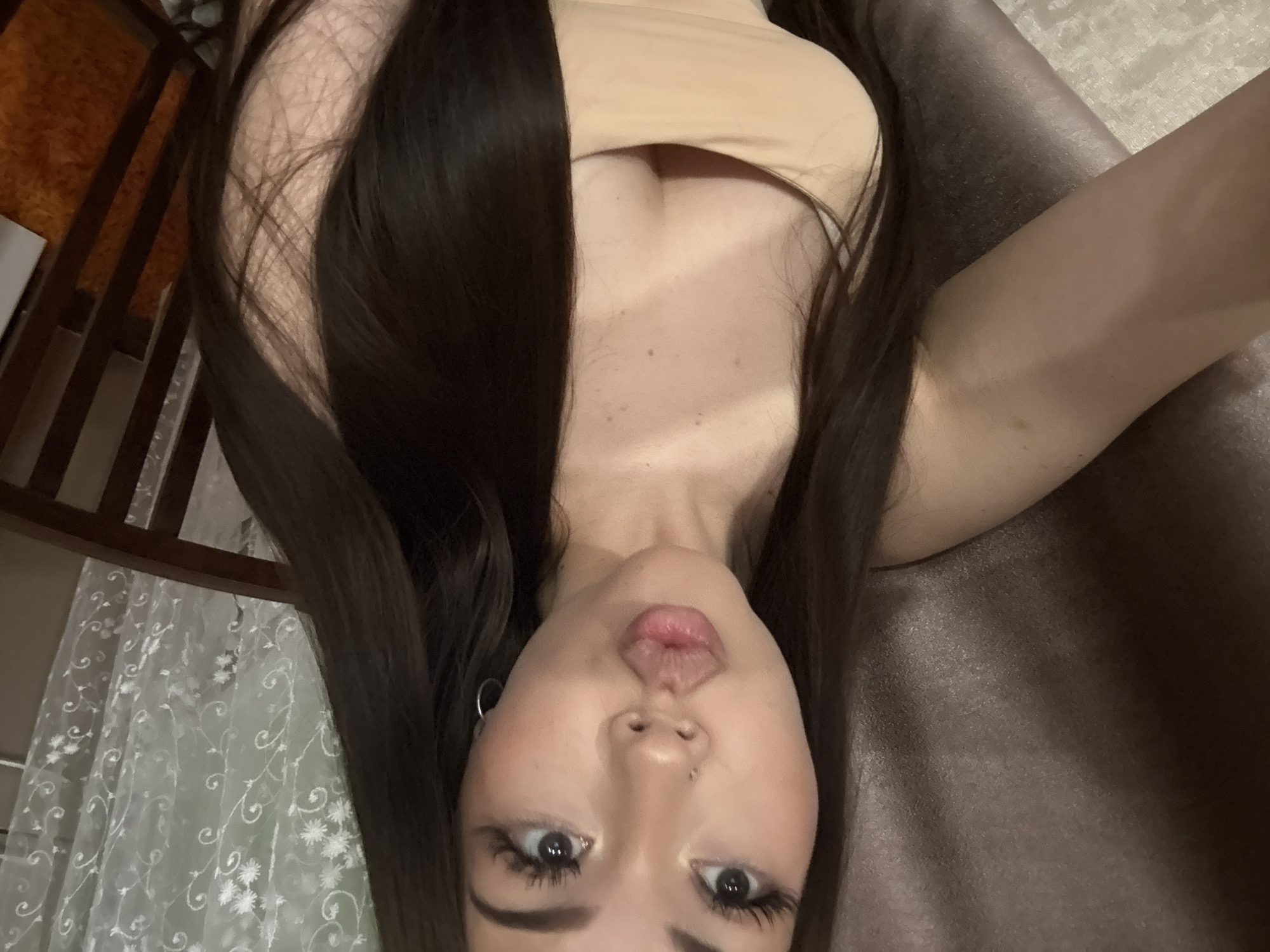 A close-up selfie captures a young woman lying down and looking upwards at the camera, giving the impression that she is upside down due to the camera's orientation. Her long, dark hair cascades around her face and shoulders. She wears a light beige or nude-colored strapless top, and faint tan lines are visible on her chest. Her lips are slightly parted in a playful expression, with her tongue visible, and her wide eyes are focused on the lens. She has dark eyeliner/mascara, defined eyebrows, a small nose piercing, and a silver hoop earring in her left ear.
The indoor setting suggests a home in Naberezhnye Chelny, Russia. To her right, a dark, textured surface, possibly a sofa or upholstered furniture, occupies the background. To her left, wooden furniture elements, possibly part of a chair or bed frame, are visible, alongside a white, sheer curtain adorned with a delicate floral pattern, suggesting a window nearby. The lighting is soft and ambient. No text is visible in the image.