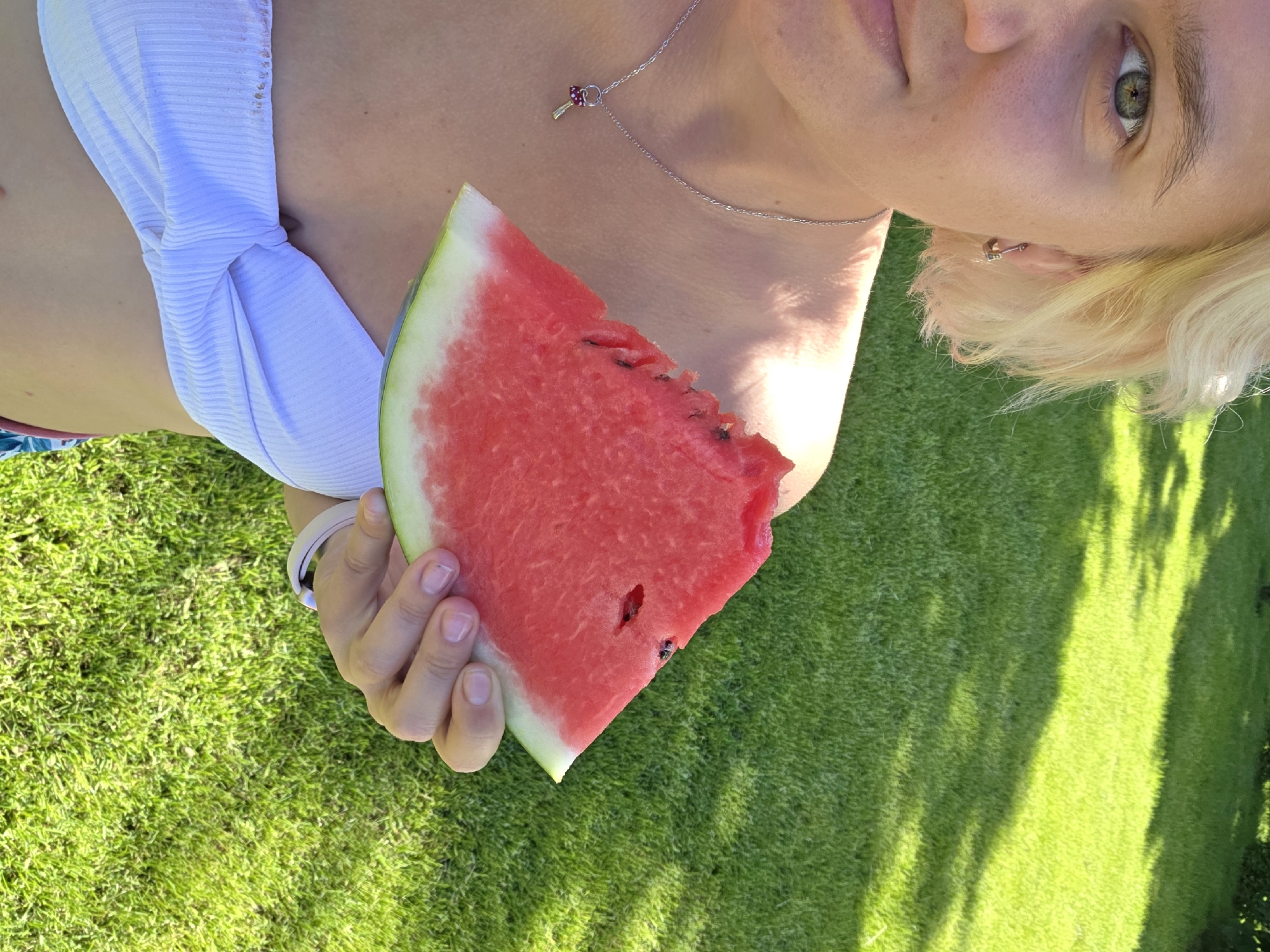 In Frolovo, Russia, on a sunny day, a person is captured from the chest up, holding a slice of watermelon in their right hand. The person has short, blonde hair and is wearing a white strapless top, a silver necklace with a small charm, and an earring in their left ear. Their left eye, which is green, is visible, and they are looking towards the camera. The background consists of a lush green lawn with shadows cast by sunlight, suggesting an outdoor setting. The image is taken outdoors, in a grassy area.