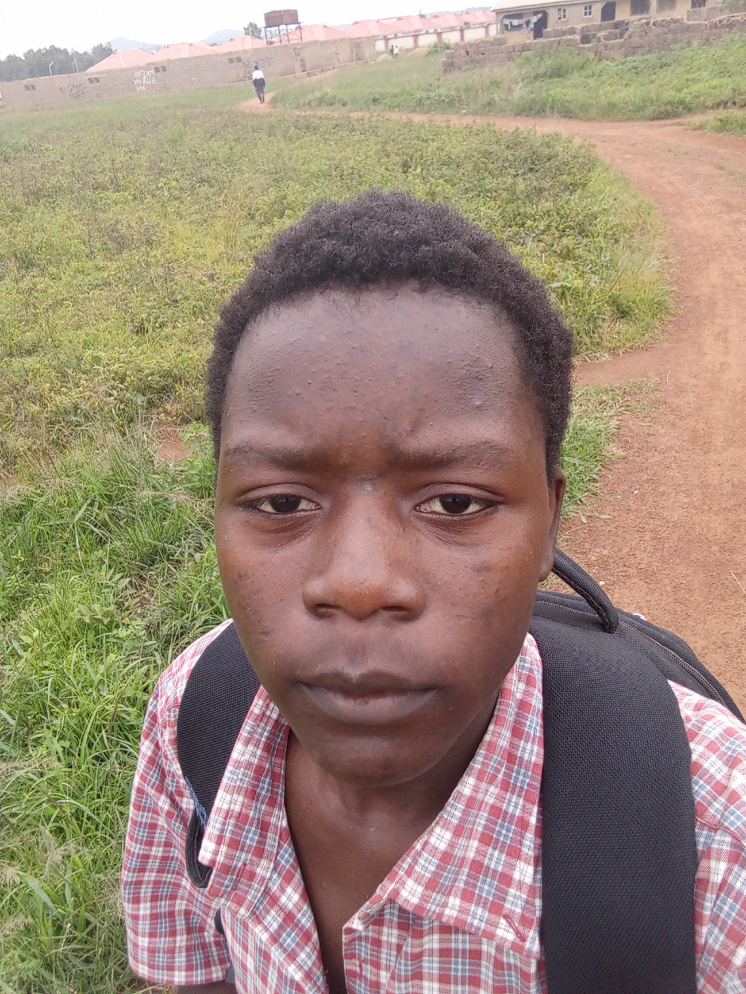 The image is a selfie taken by a young person in an outdoor setting. The subject is a person with dark skin and short, dark, curly hair, wearing a red and white plaid shirt and a black backpack. They are looking directly at the camera with a neutral expression. The background consists of a dirt path and an area of overgrown green grass and vegetation. In the distance, there are some buildings with red roofs, possibly part of a school or residential area. A water tank is visible on one of the buildings. There is also a person walking away from the camera in the middle distance, on the dirt path. The overall impression is of a bright, overcast day in a rural or semi-rural location. There is no visible text in the image. The location context is Durimi, Nigeria.