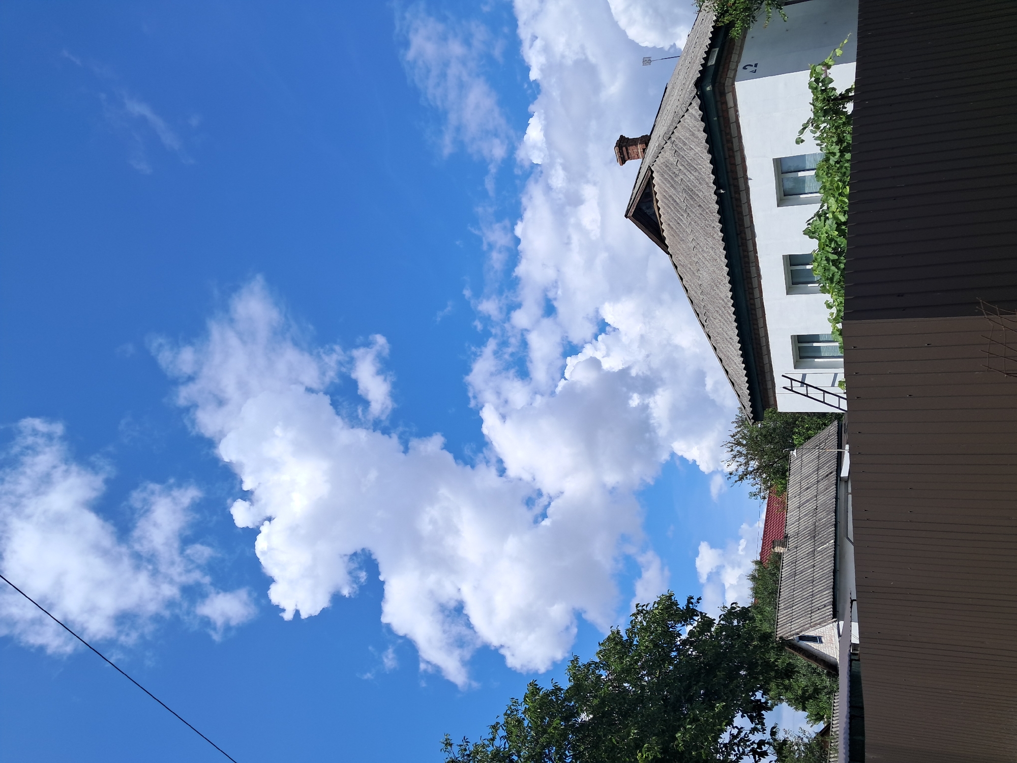 The image depicts a residential area under a bright blue sky with scattered white clouds. In the foreground, a dark brown metal fence runs horizontally across the frame. Behind the fence, to the right, a white house with a grey corrugated roof is visible. The house has three windows and a brick chimney on the roof. A ladder leans against the right side of the house. To the left of this house, another building with a similar grey roof is partially visible. Lush green trees border the properties. The image was taken in the city of Kremenchuk, Ukraine.