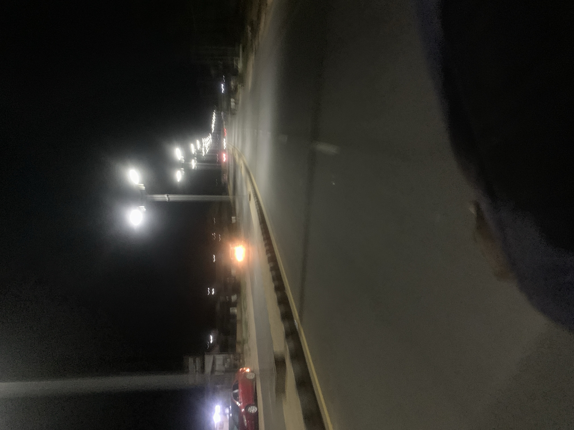 A nighttime image shows a street in Adankolo, Nigeria, with streetlights illuminating a dark road and a red car parked on the side. The image is taken from an elevated position, looking down the road.