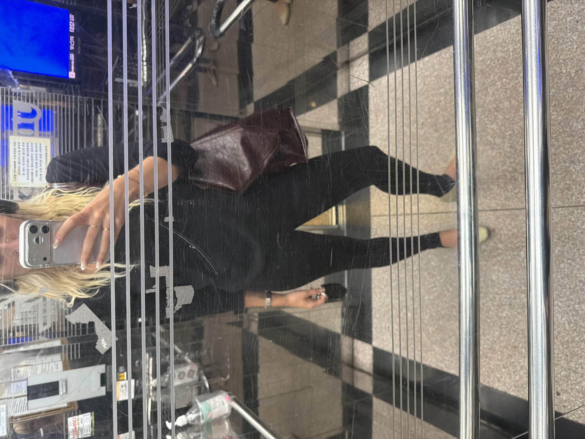 A woman is taking a selfie in an elevator with a mirrored wall. She is wearing a black top and leggings and holding a silver smartphone in her left hand. A brown leather handbag is slung over her shoulder. The elevator has a checkered floor and chrome handrails. Inside the elevator, there is a display screen showing advertisements in Korean and English. The elevator is likely in Seoul, South Korea, based on the language on the display screen.