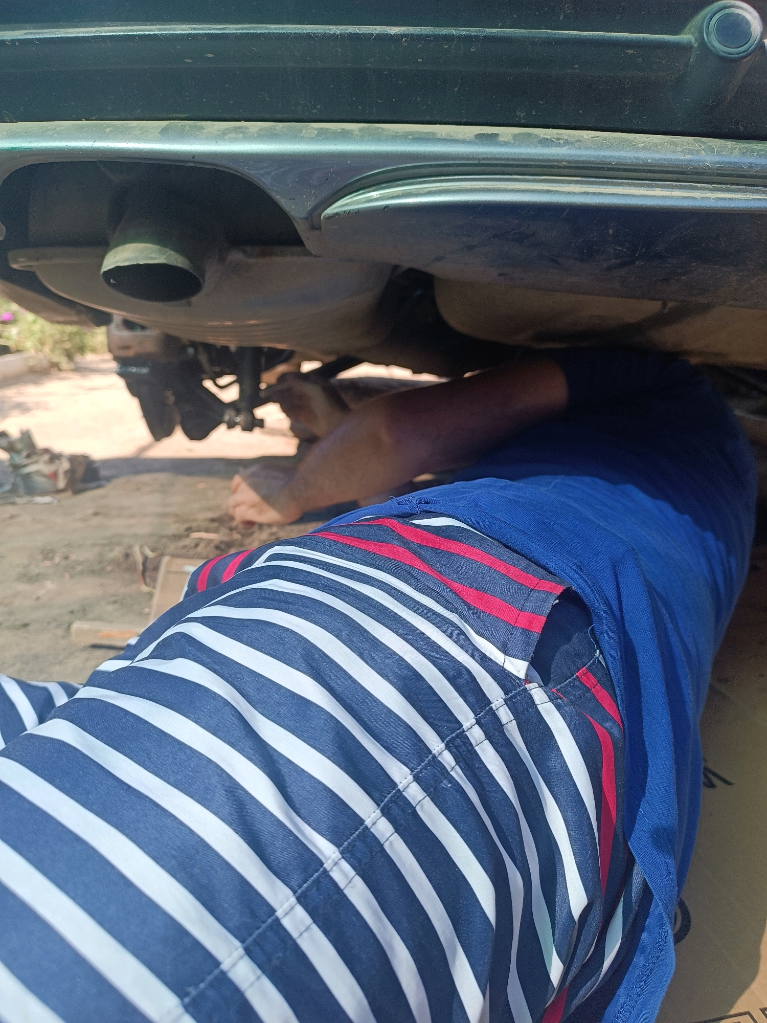 In Vypasne, Ukraine, a person is lying on the ground, working underneath a vehicle. They are wearing striped pants and a blue shirt. The vehicle appears to be a car, with its underside and exhaust system visible. The ground is dusty and uneven, with some debris scattered around. The scene is outdoors, with sunlight illuminating the area.