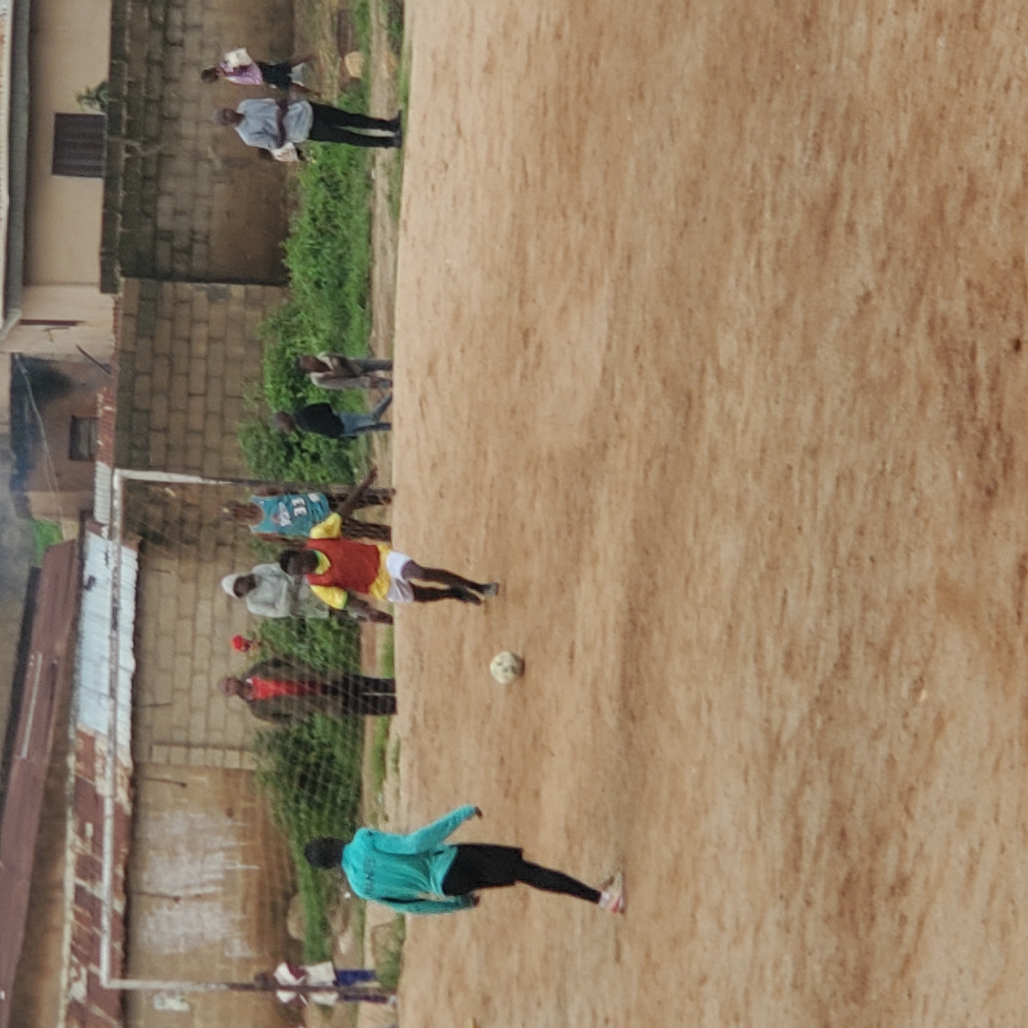 In an outdoor, dusty, and undeveloped football field, several individuals are present. A young person in a bright blue shirt and black shorts, with black socks, is in motion towards the left of the frame, facing away from the camera. Another person, wearing a yellow shirt and red vest over white shorts and black socks, is actively playing football, with their right leg extended towards the ball, which is visible on the ground in front of them.
In the background, a weathered brick wall with sections of corrugated metal roofing forms a backdrop. A goalpost with netting is visible on the left side of the frame. Several adults and children are scattered across the background, some standing and observing, others in closer proximity to the goal. One adult in a light blue shirt and black pants stands in the middle ground, appearing to be watching the game. A person in a pink top and black shorts walks across the far right. Further to the left, a person in a red jacket stands with their back to the camera. Other figures, including children, are less distinct. Green foliage borders the area behind the wall. The ground of the football field is a light brown, sandy dirt.