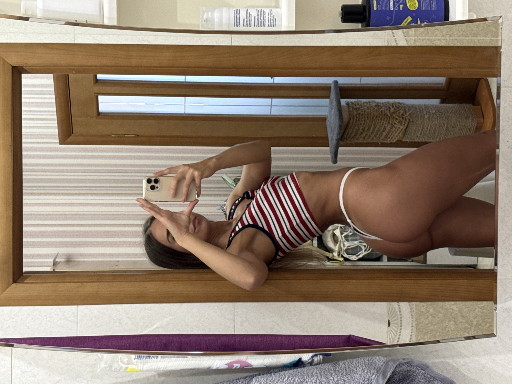 A young woman with long brown hair is taking a mirror selfie in what appears to be a bathroom. She is wearing a red and white striped crop top and white thong. She is leaning against a wooden door frame and her left hand is raised, holding a smartphone. The background shows a wall with vertical striped wallpaper, a tiled wall, and shelves with various bathroom products. A cat scratching post is also visible. There are no explicit indications of the location being in Saint Petersburg, Russia, other than what was stated in the prompt.