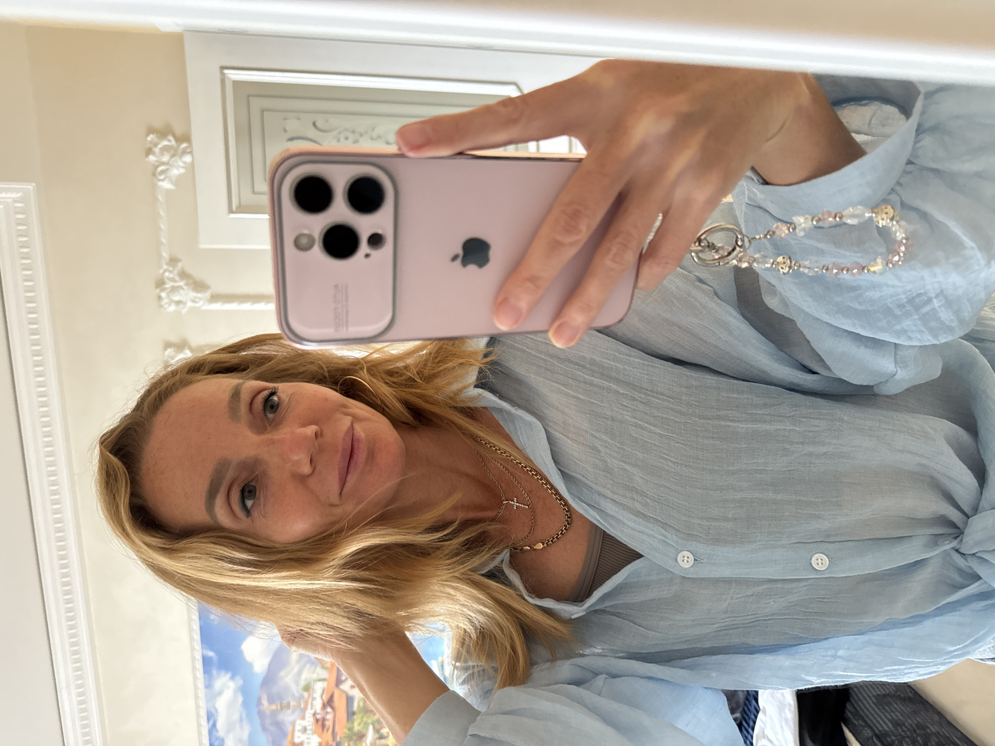 A woman is taking a mirror selfie in what appears to be a well-decorated room. She has blonde hair and is wearing a light blue, slightly wrinkled button-down shirt with a knot in the front. She is also wearing necklaces, one of which has a cross pendant. Her phone, a pink iPhone, is held in her right hand. A beaded keychain is attached to the phone. The background shows a mirror reflecting a painting of a European-style town with mountains and a blue sky. The room also features ornate white moldings on the wall and ceiling. The overall impression is one of a casual, indoor setting. The location is unknown.