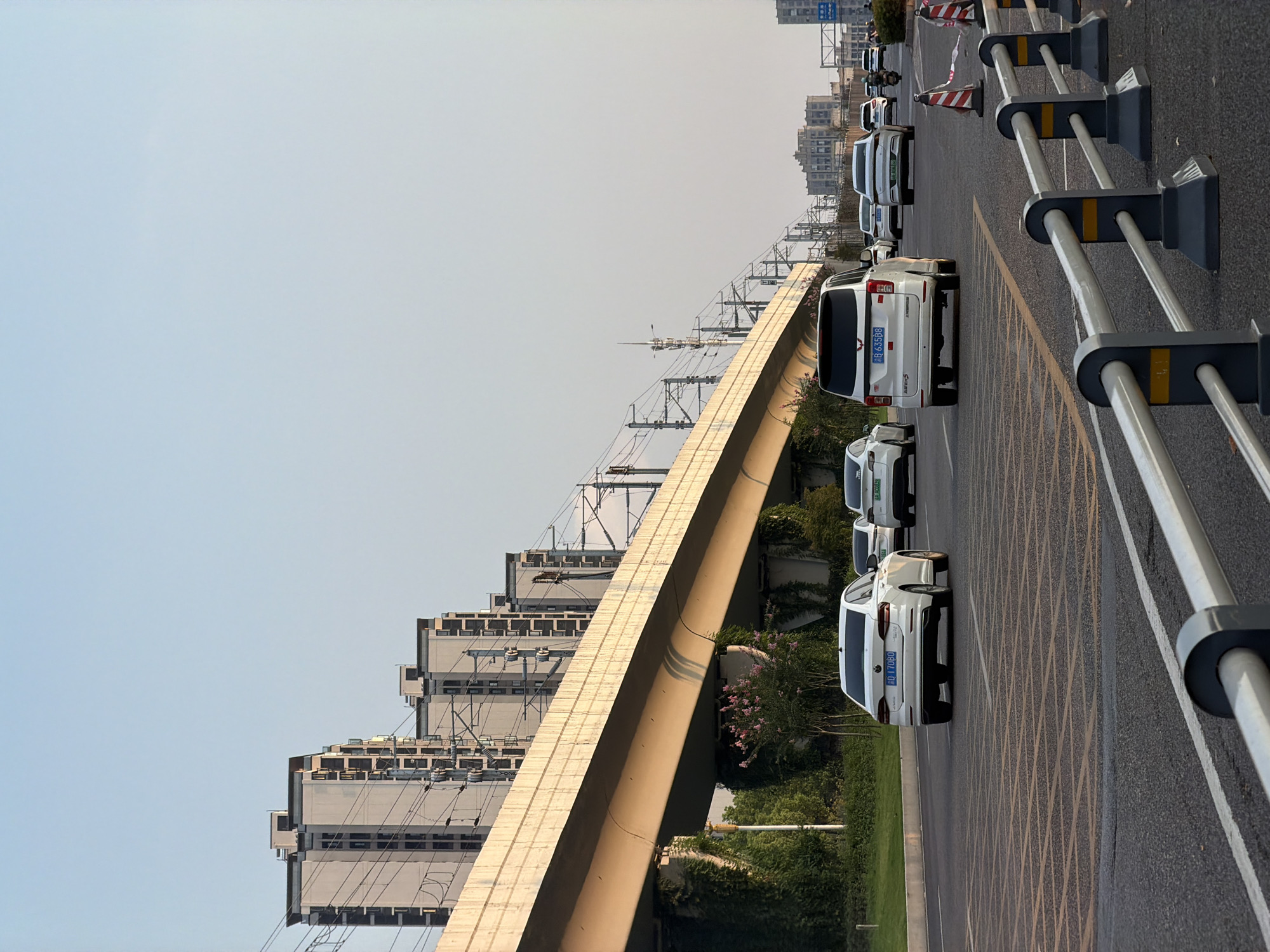 In Jiangjin, China, an overpass for a light rail system runs diagonally across the frame from the left foreground to the right background, casting shadows on the road below. Several white vehicles are visible on the multi-lane road, with a white van in the middle lane and white sedans in adjacent lanes, all heading towards the right. On the left side of the road, a grassy embankment is covered in ivy, with a flowering bush in bloom. Behind the overpass, a row of modern, light-colored apartment buildings with dark-framed windows is visible, stacked in a staggered pattern. The sky is clear and a pale blue. Safety barriers with red and white striped cones are positioned along the right side of the road in the foreground.