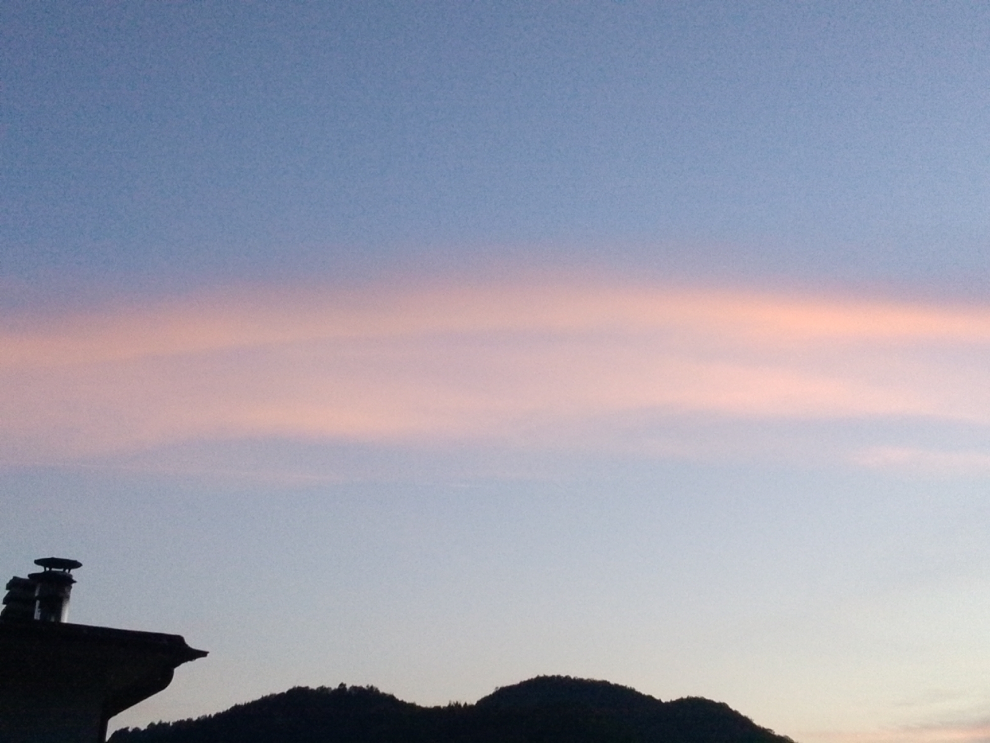 The image captures a serene twilight scene over Valle Castellana, Italy. A silhouette of a house with a chimney stands against the fading light on the left side, while the dark outline of a forested hill stretches across the lower right portion of the frame. The sky is a gradient of soft blue and pink hues, with a delicate band of pink clouds stretched horizontally across the middle. The overall atmosphere is tranquil and picturesque, indicative of a peaceful evening.