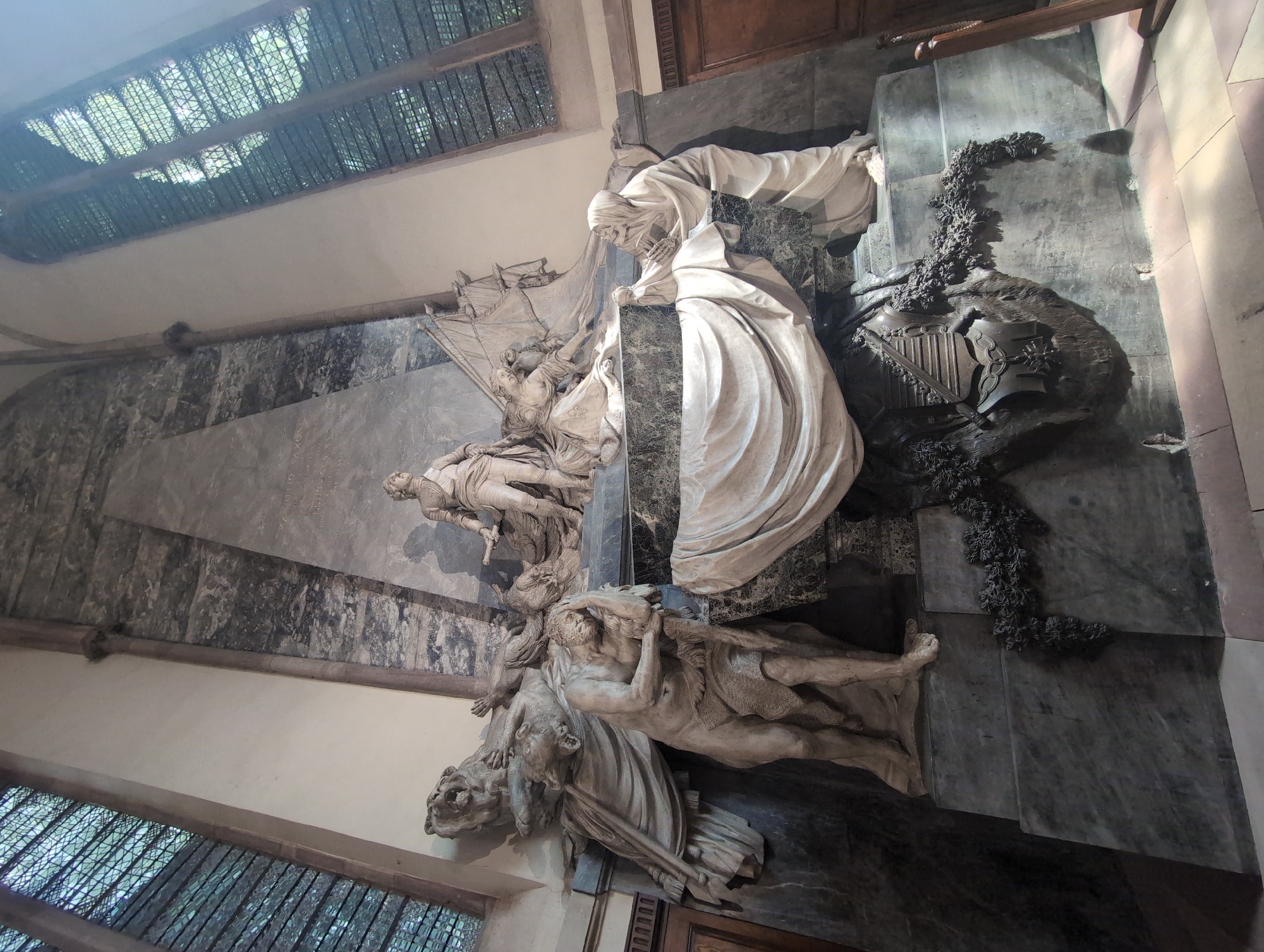 This is a photograph of the monument to Louis XV in Strasbourg Cathedral, France. The monument is a large, marble sculpture depicting several figures. In the center, a standing male figure in military attire is depicted. To the left, a muscular male figure with a pained expression is shown, leaning on a staff, with a lion and a bird at his feet. To the right, a female figure kneels, reaching towards the central figure, and another figure, resembling Death with a skull and a book, is seated. The monument is set against a large marble slab with inscriptions, and is surrounded by draped fabric and decorative elements. Tall, arched windows with ornate grilles are visible in the background, suggesting an interior religious setting. The lighting is dramatic, with sunlight streaming through the windows, creating shadows and highlighting the sculpted details.