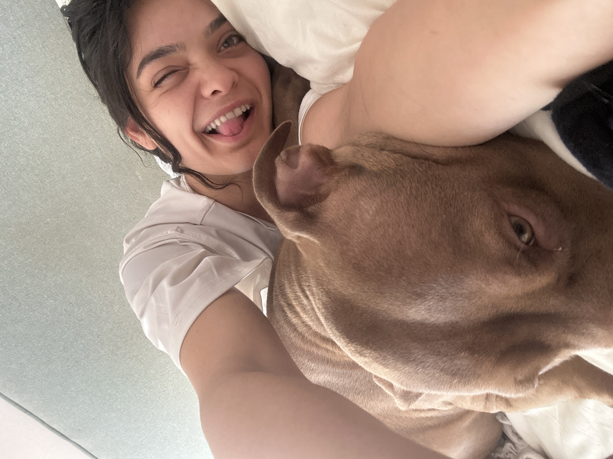 A person and a dog are lying on a bed. The person is smiling and sticking their tongue out at the camera. The dog's head is resting on the person's arm. The room appears to be a bedroom, with light-colored bedding and a grey wall. No specific location within Malevo, Bulgaria is identifiable.