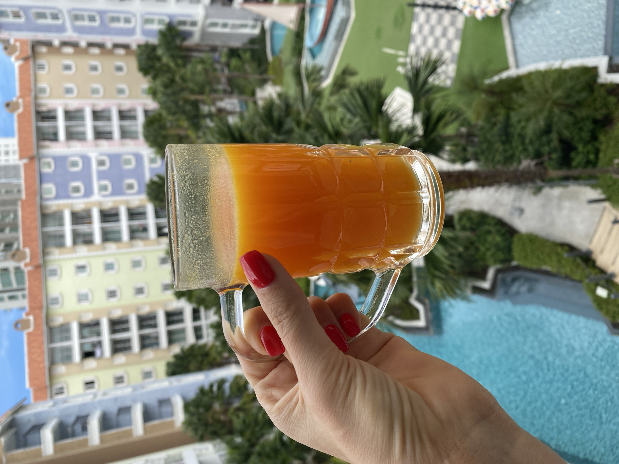 A close-up shot shows a person's hand with bright red nail polish holding a clear glass mug filled with orange juice. The mug has a textured pattern. The background is blurred but depicts a resort or hotel complex in Pattaya City, Thailand, with colorful buildings, palm trees, and swimming pools. The sky is visible and appears partly cloudy.